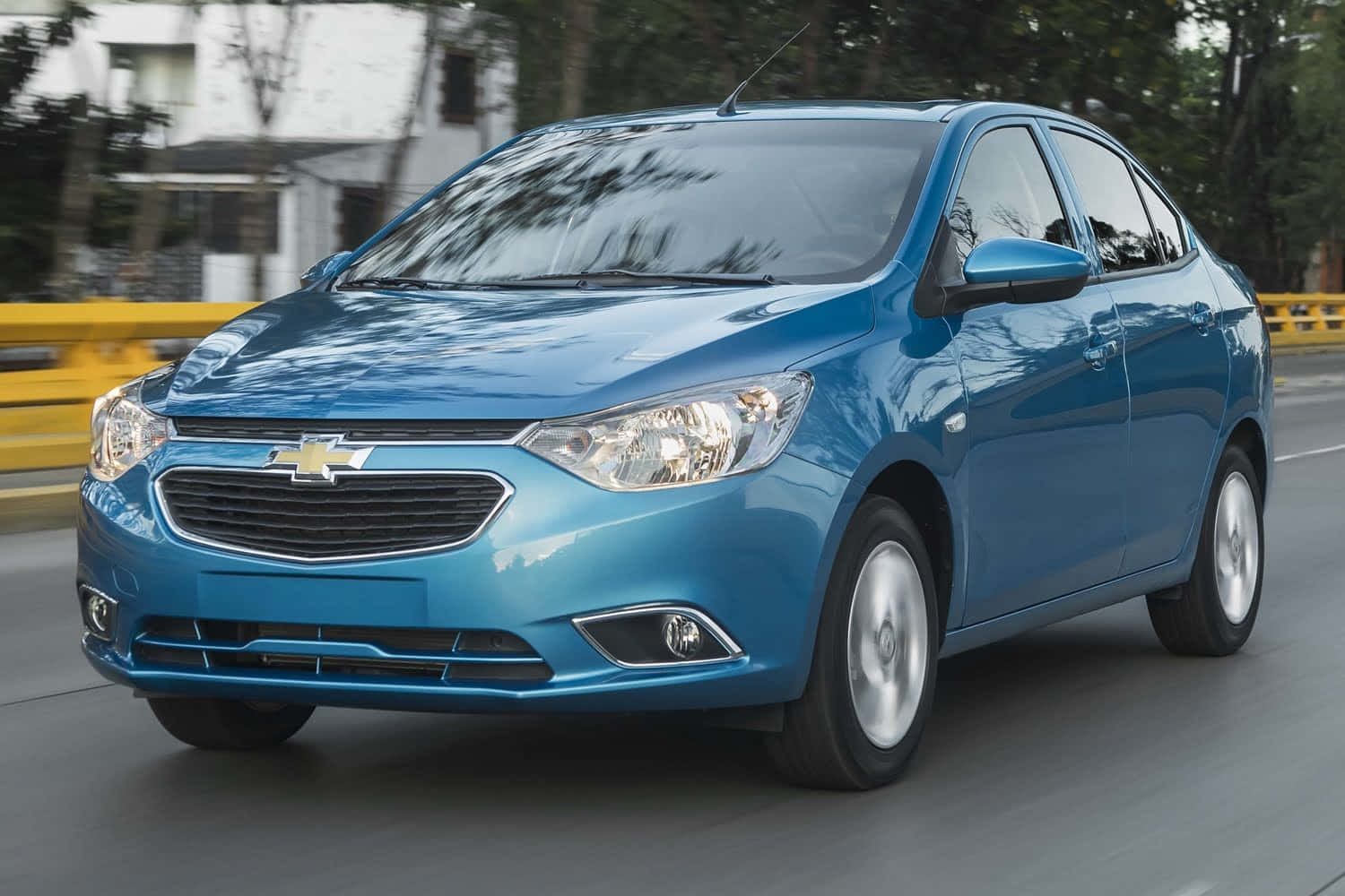 Chevrolet 桌布 壁紙 — 一輛充滿活力的藍色雪佛蘭轎車在鋪砌的道路上行駛，背景是模糊的樹木和建築物。