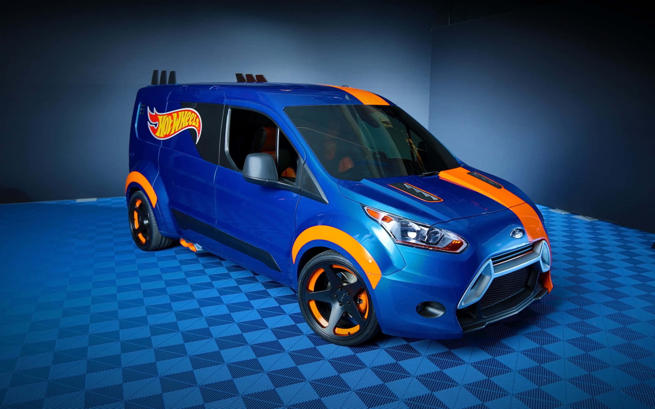 Ford Transit 桌布 壁紙 — 一輛充滿活力的藍色風火輪廂型車，帶有橙色點綴和定制輪圈，在工作室燈光下停放在格子地板上。