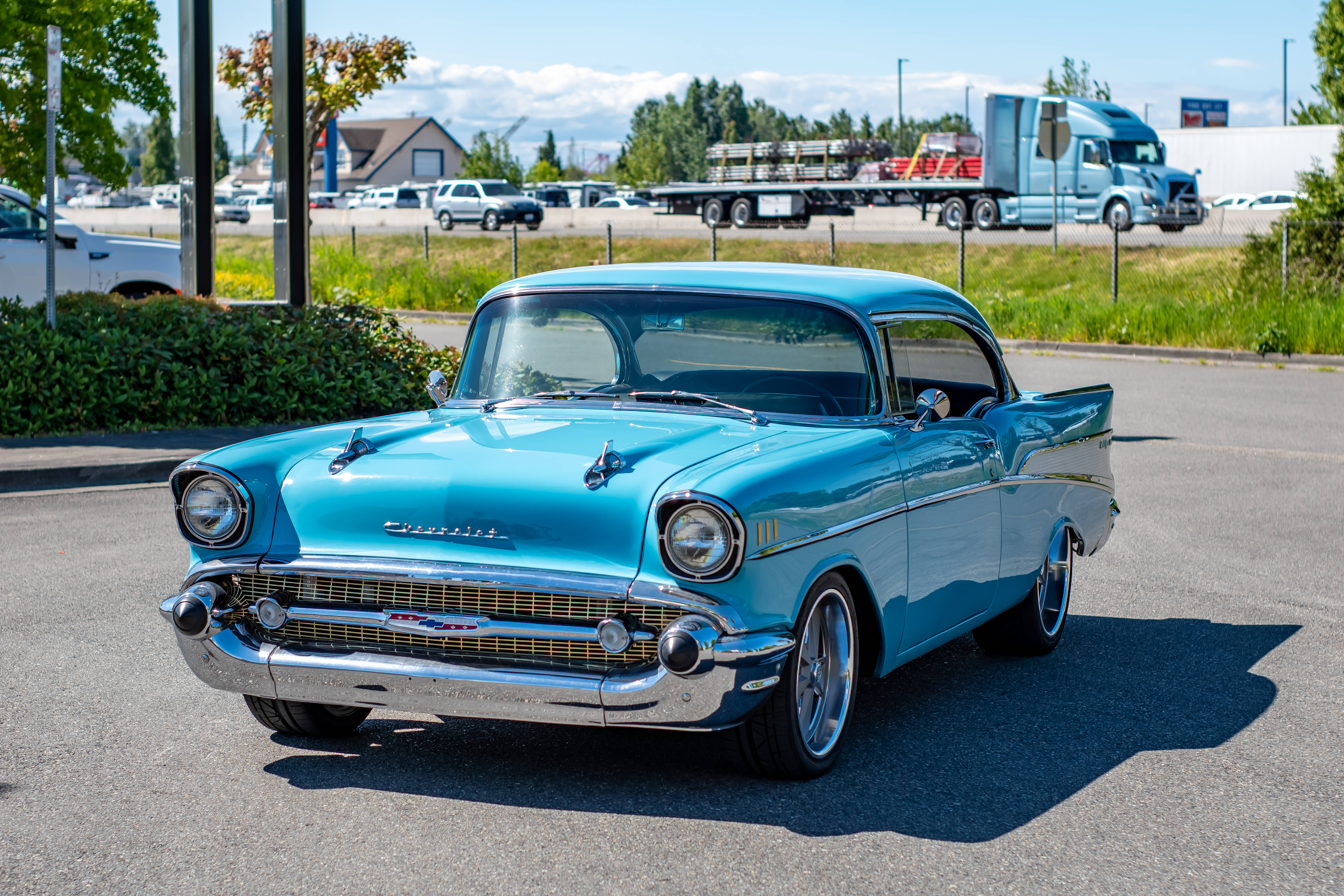Chevrolet 桌布 壁紙 — 一輛充滿活力的青綠色1957年雪佛蘭Bel Air在明亮的陽光下，在鋪砌的停車場中閃閃發光，背景有卡車和樹木。