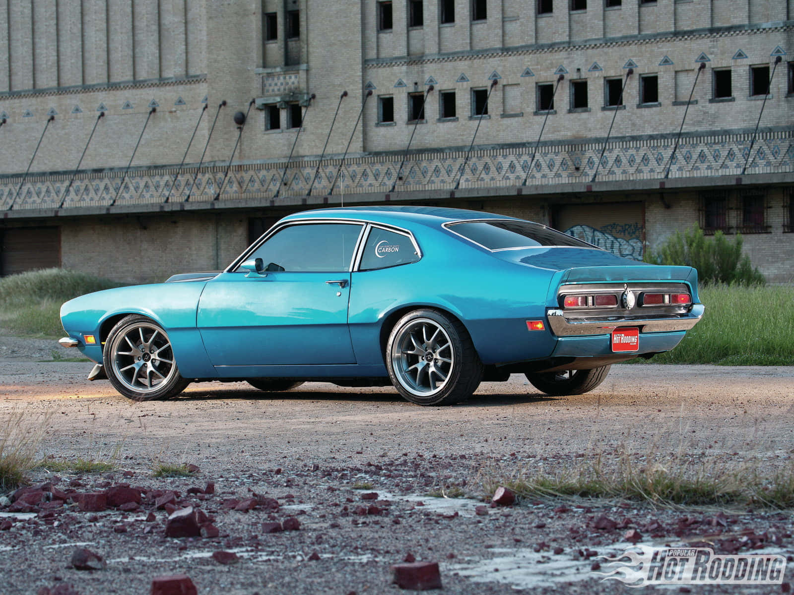 Ford Maverick 桌布 壁紙 — 一輛充滿活力的青綠色經典肌肉車停在碎石地上，前面是一座風化的工業建築。