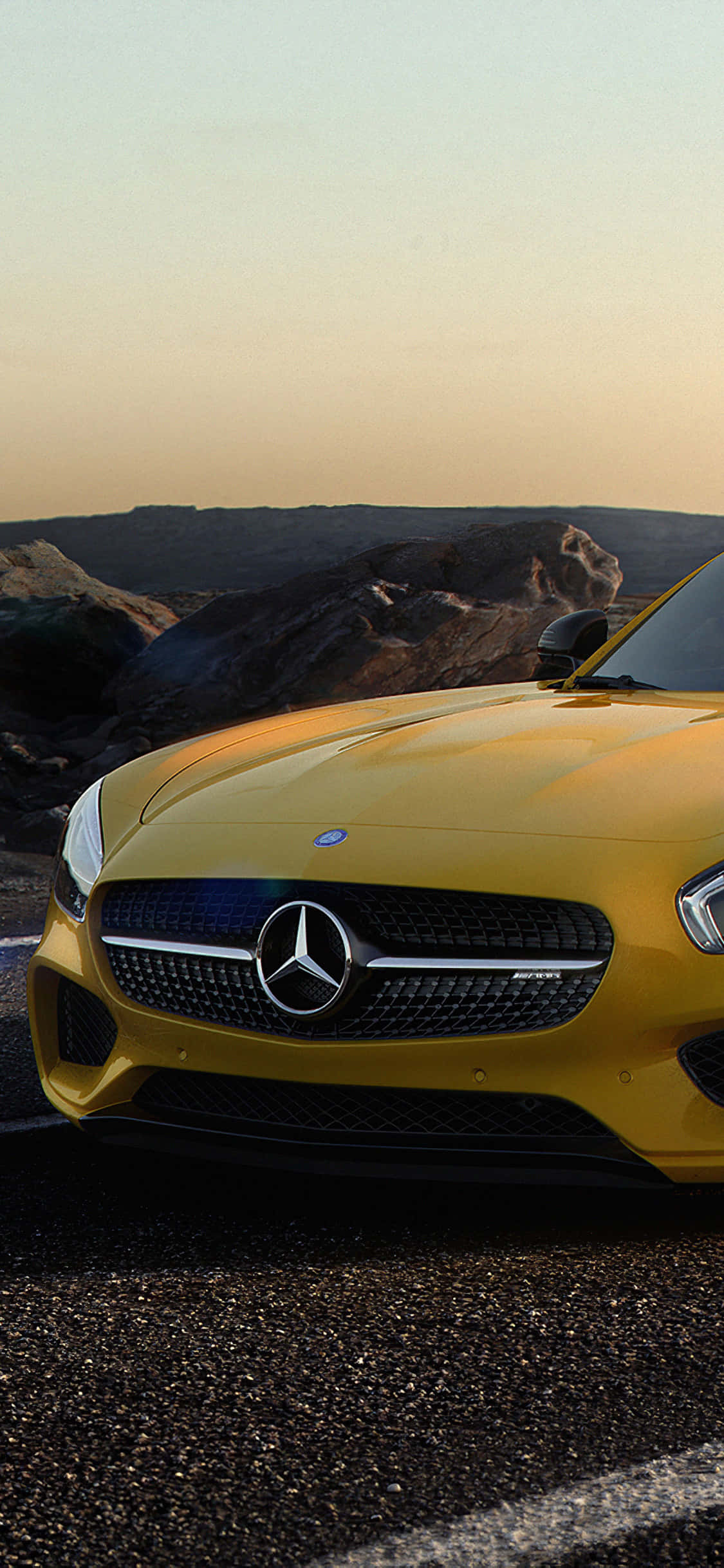 iPhone X Amg Gt-r 桌布 壁紙 — 一輛充滿活力的黃色 Mercedes-AMG 跑車在金色夕陽下閃閃發光，停在碎石路上，旁邊是崎嶇的沙漠山丘。
