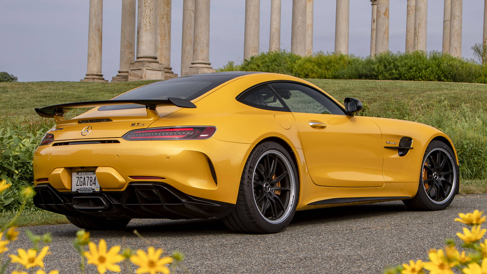 Gtr 桌布 壁紙 — 一輛充滿活力的黃色 Mercedes-AMG GT R 跑車停放在碎石路上，前景有古老的石柱和黃色花朵。