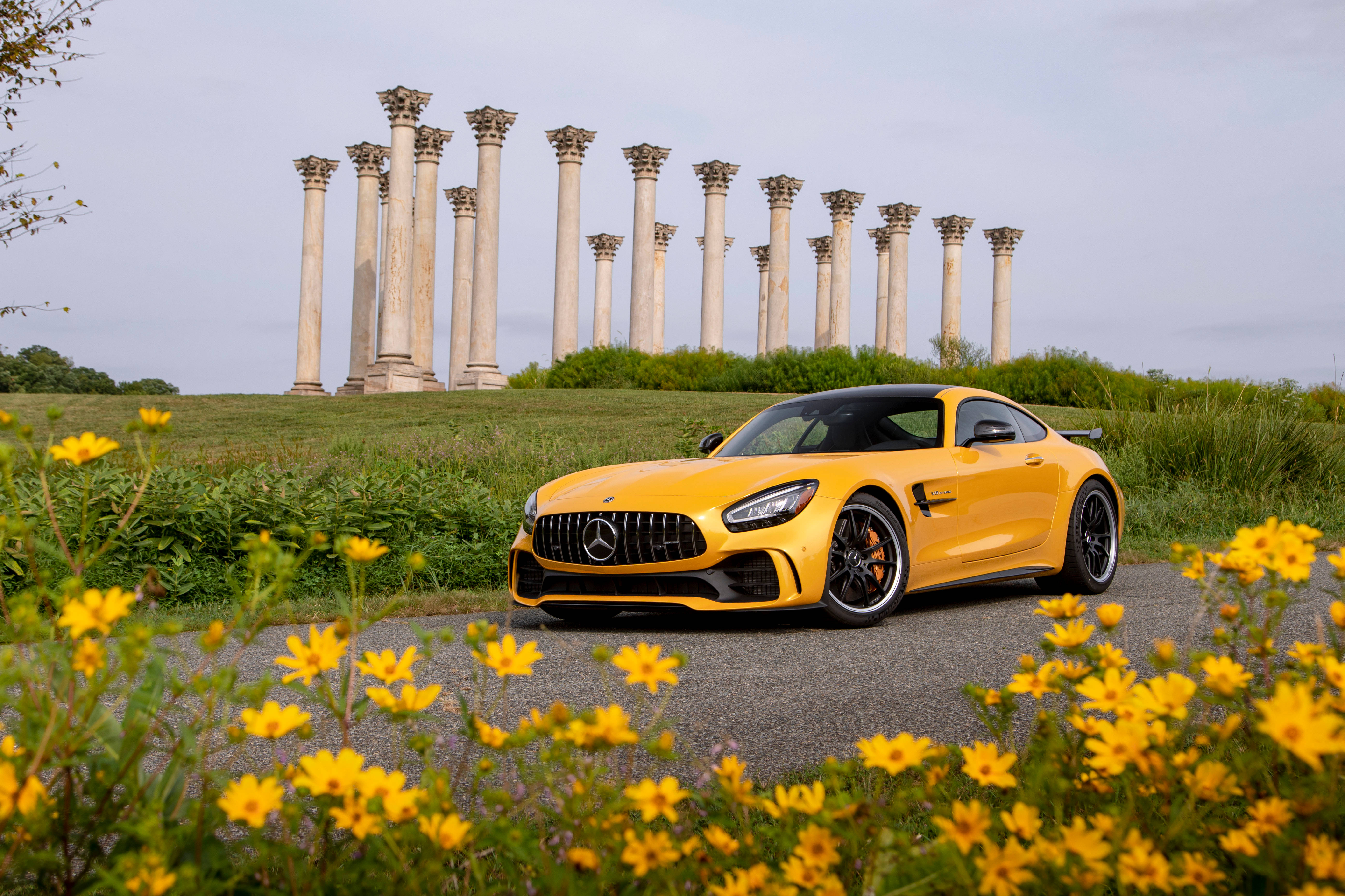 R 桌布 壁紙 — 一輛充滿活力的黃色 Mercedes-AMG GT 停泊在碎石路上，兩旁是黃色野花，背後草坡上矗立著古老的石柱。