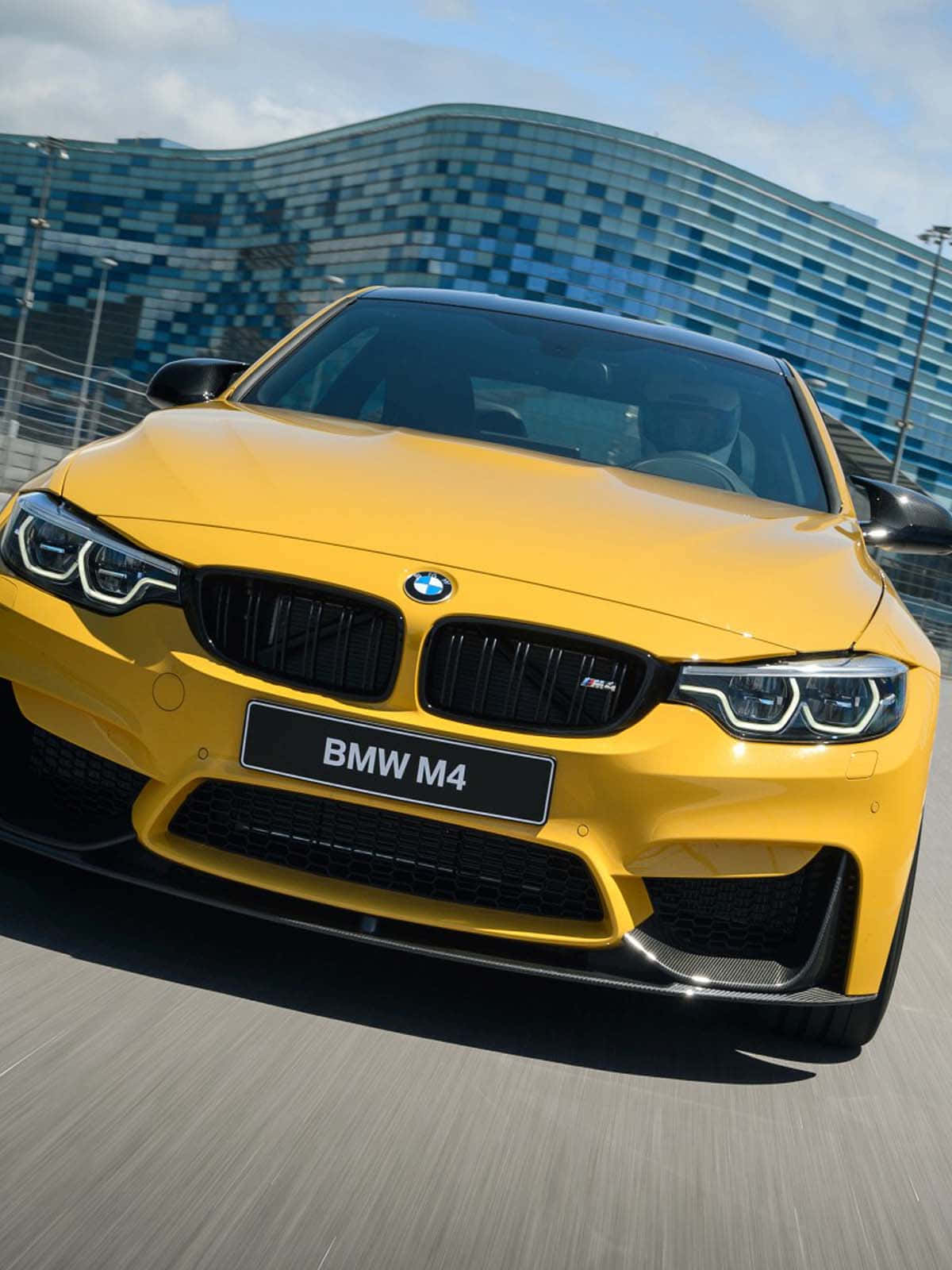 BMW 動圖 桌布 壁紙 — 一輛充滿活力的黃色BMW M4跑車在晴朗的天空下，以動態模糊效果飛馳而過一座現代玻璃建築。