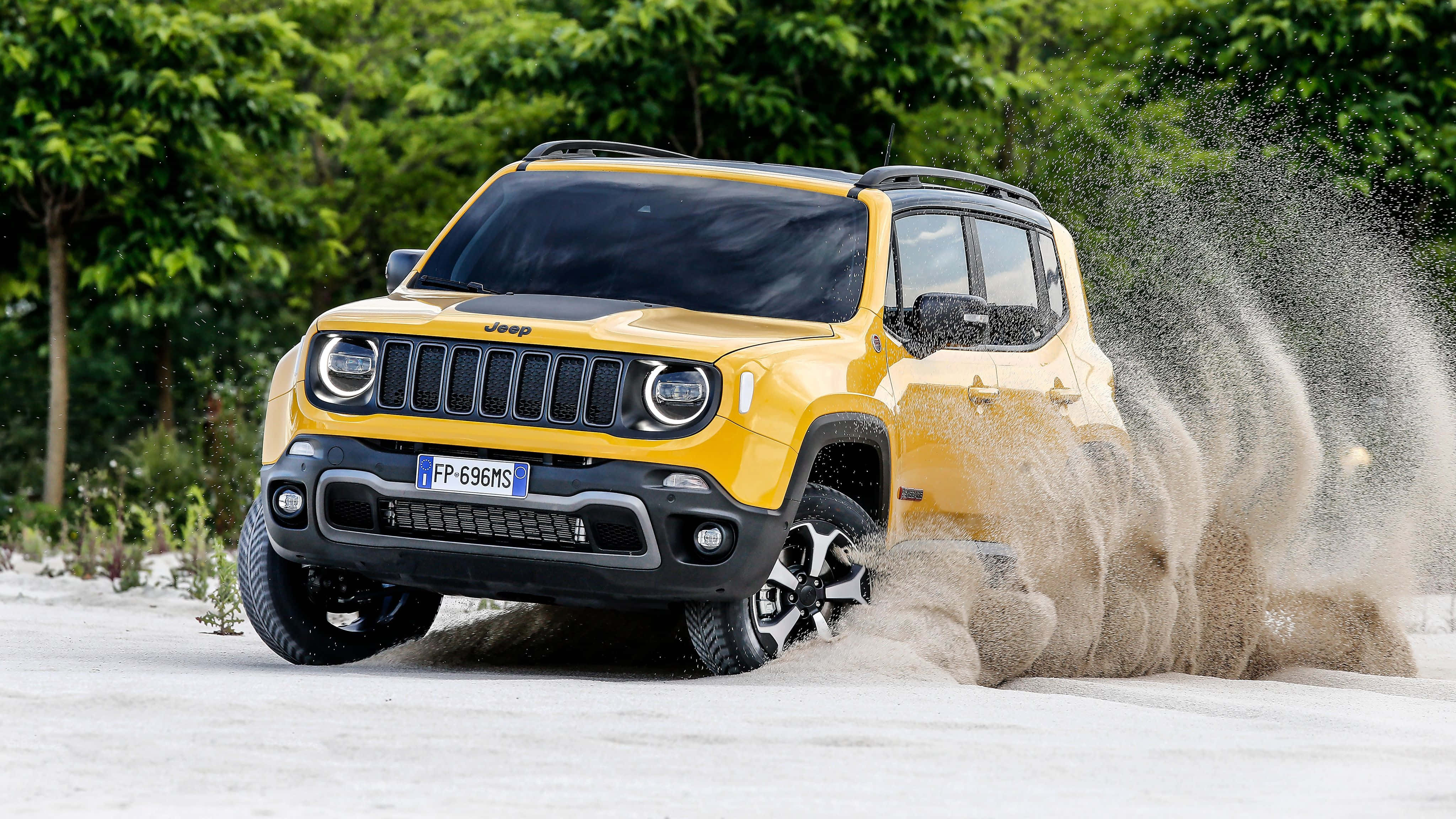 Jeep Renegade 桌布 壁紙 — 一輛充滿活力的黃色Jeep Renegade在沙地中強勁行駛，揚起戲劇性的塵埃雲，背景是茂密的綠樹。