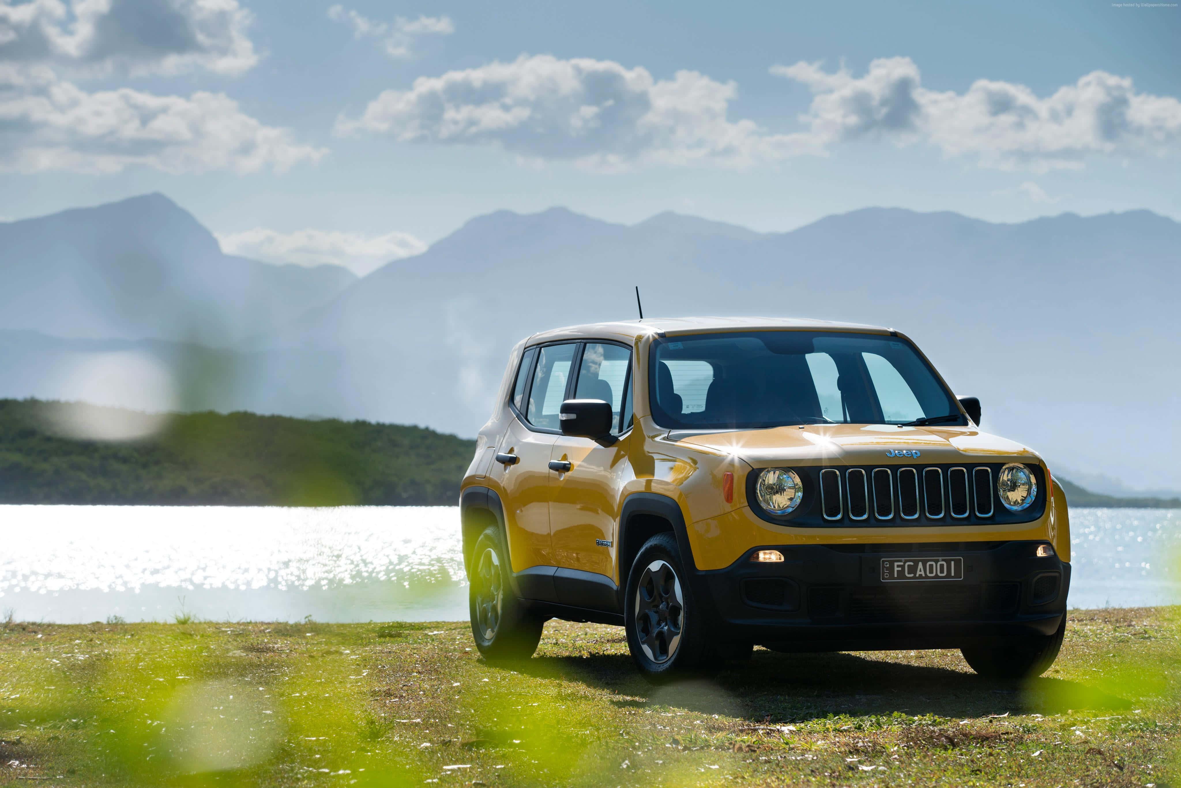 Jeep Renegade 桌布 壁紙 — 一輛充滿活力的黃色Jeep Renegade停泊在草地旁，靠近閃閃發光的湖泊，遠山在部分多雲的天空下。