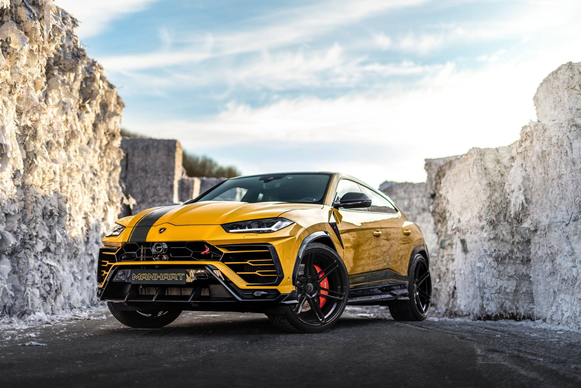 lambo 桌布 壁紙 — 一輛充滿活力的黃色Manhart Lamborghini Urus，帶有黑色飾件和紅色制動卡鉗，停放在高聳的白色石灰岩懸崖之間，天空部分多雲。