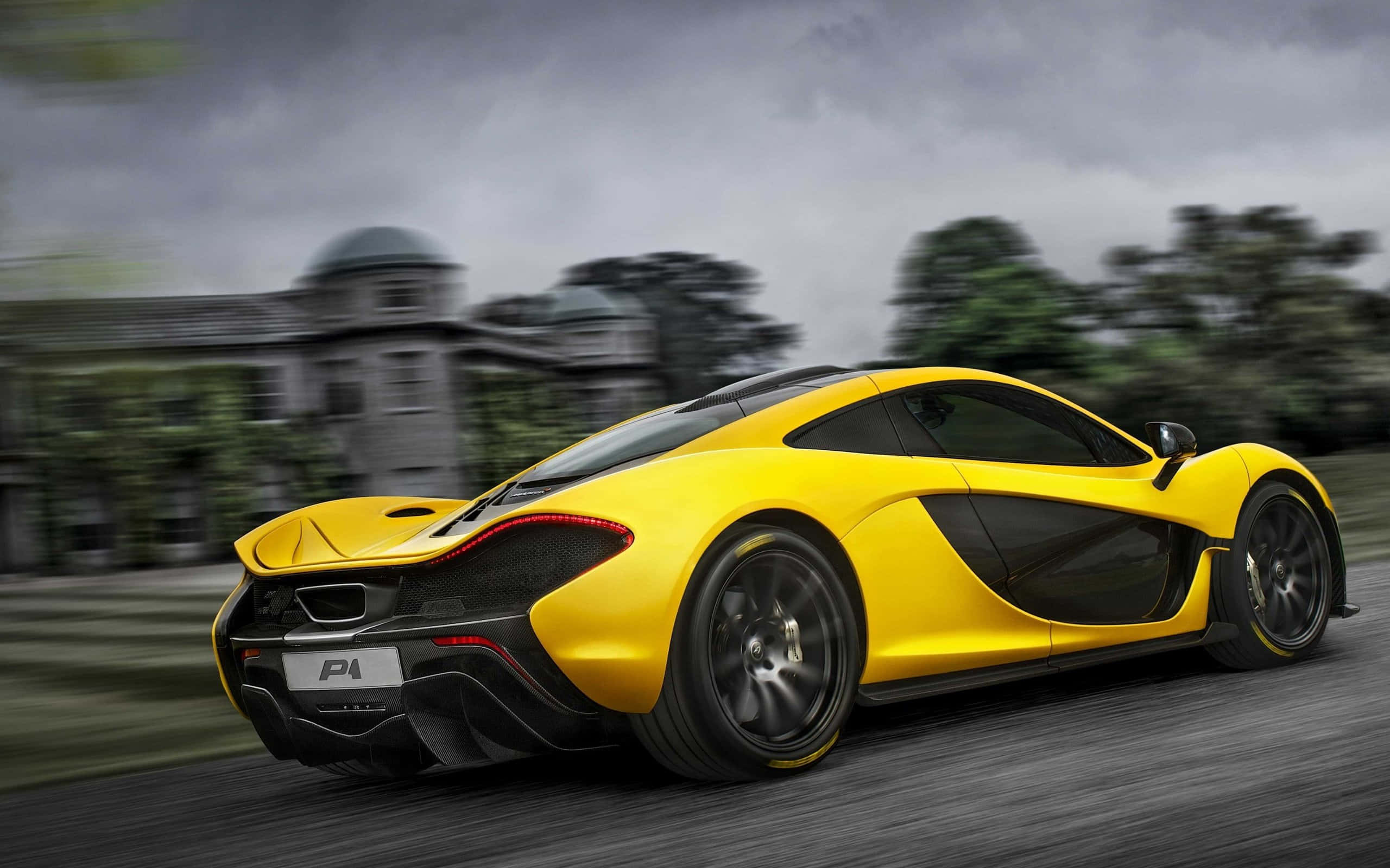 Live Car 桌布 壁紙 — 一輛充滿活力的黃色McLaren P1超級跑車在多雲的天空下飛馳經過一座宏偉的莊園。