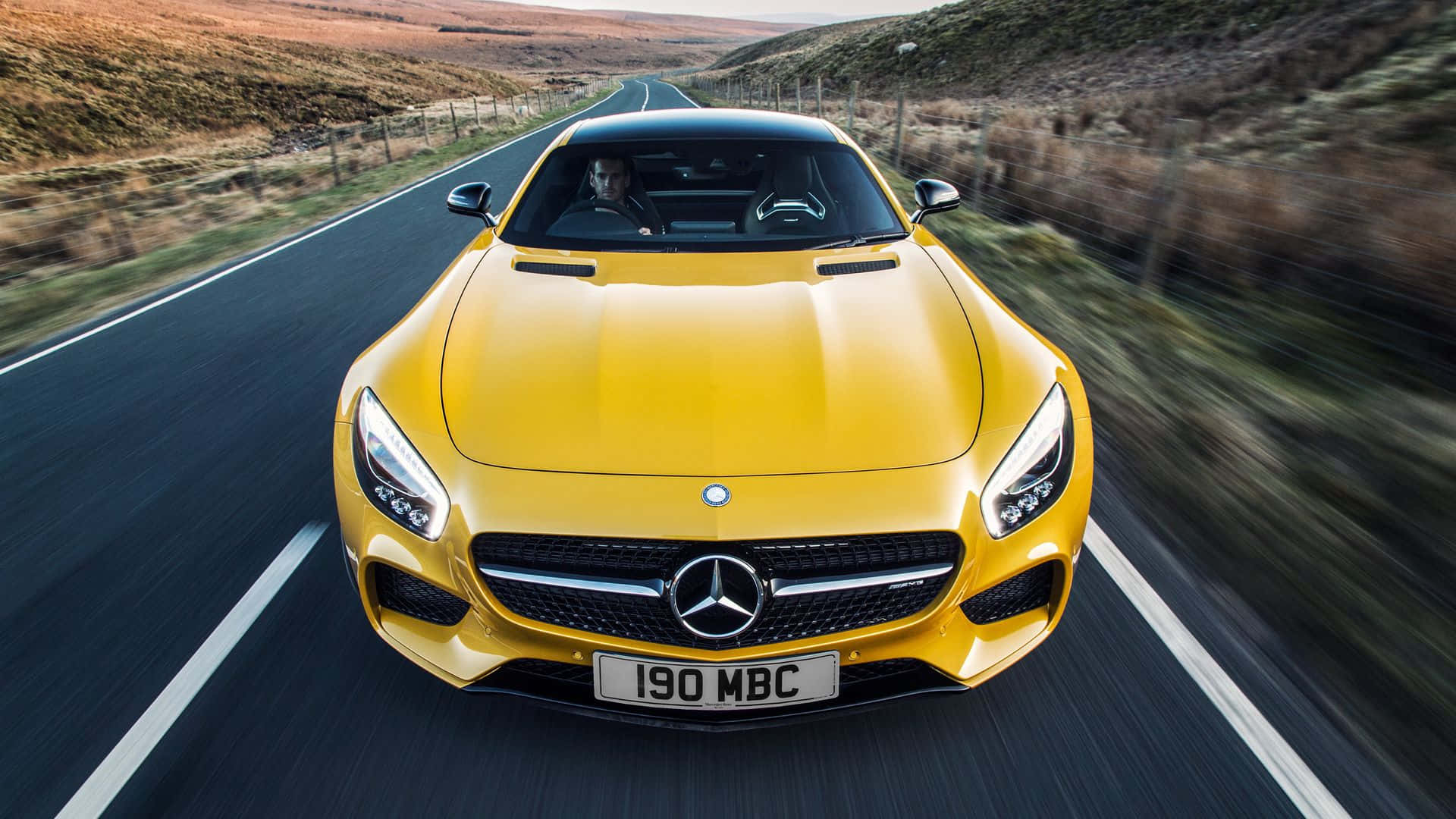 高清 桌布 壁紙 — 一輛充滿活力的黃色Mercedes-AMG GT在蜿蜒的鄉村道路上疾馳，帶有模糊的動態和金色的日落光線。