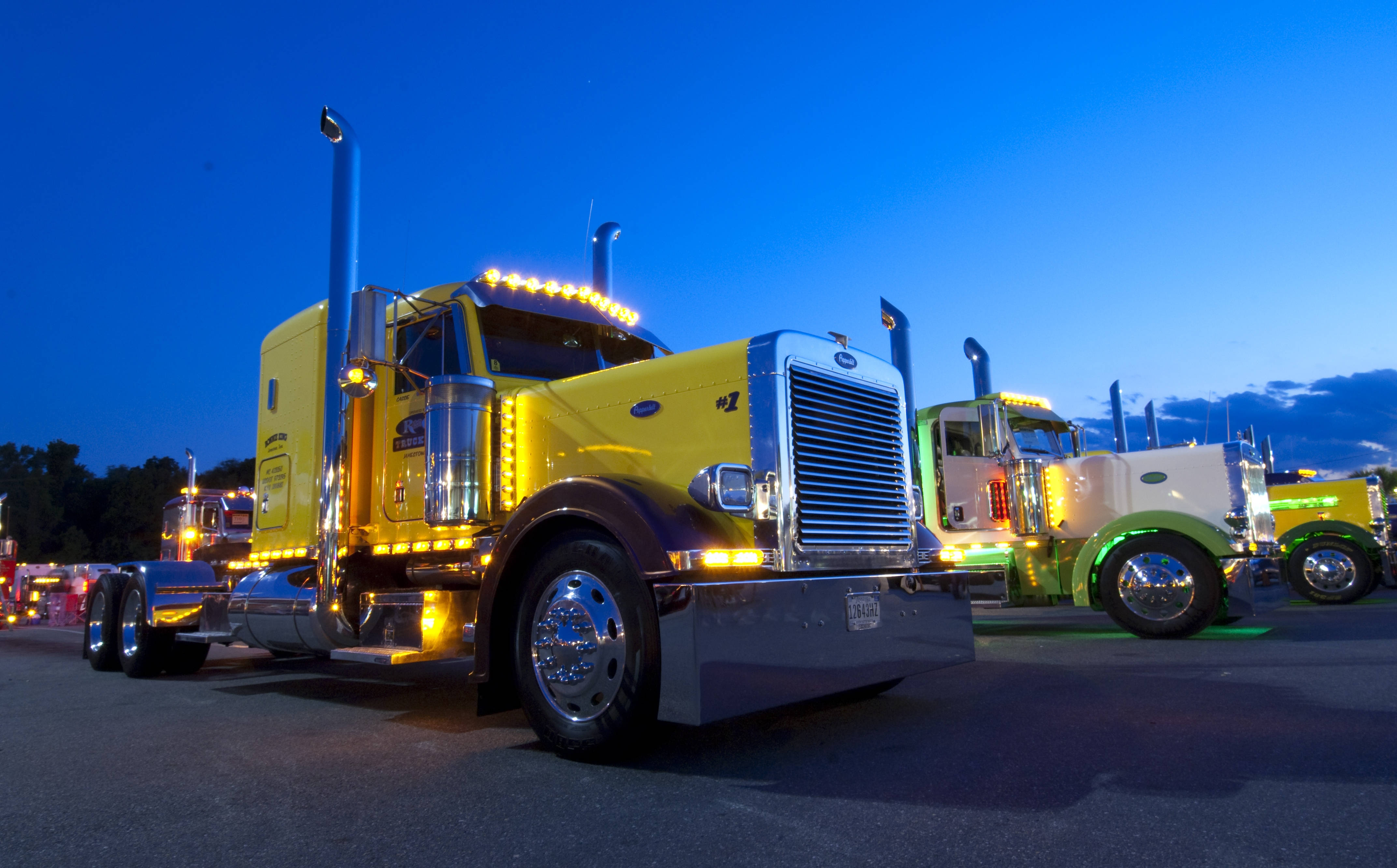 Peterbilt 桌布 壁紙 — 一輛充滿活力的黃色半掛卡車在黃昏時分閃耀著燈光，在深藍色的天空下與其他色彩繽紛的卡車並排。