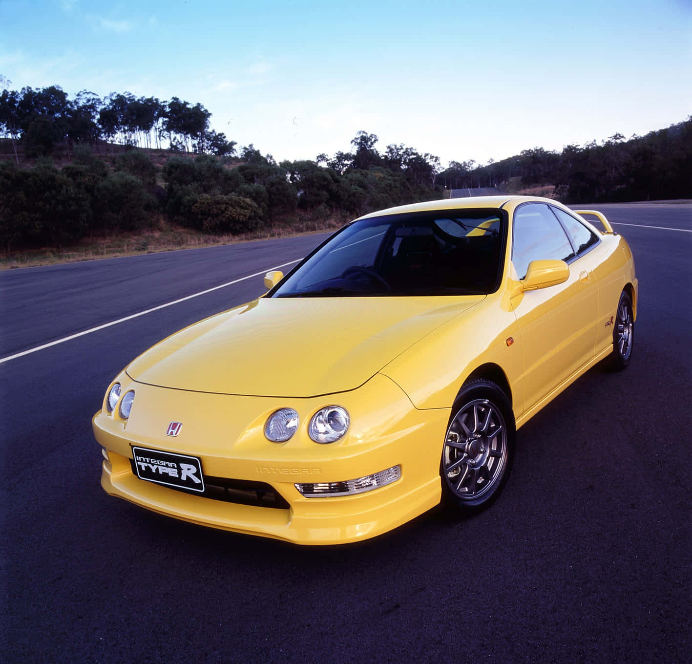 Honda Integra 桌布 壁紙 — 一輛充滿活力的黃色本田Integra Type R跑車停泊在空曠的瀝青路上，背景有樹木和山丘，天空清澈湛藍。