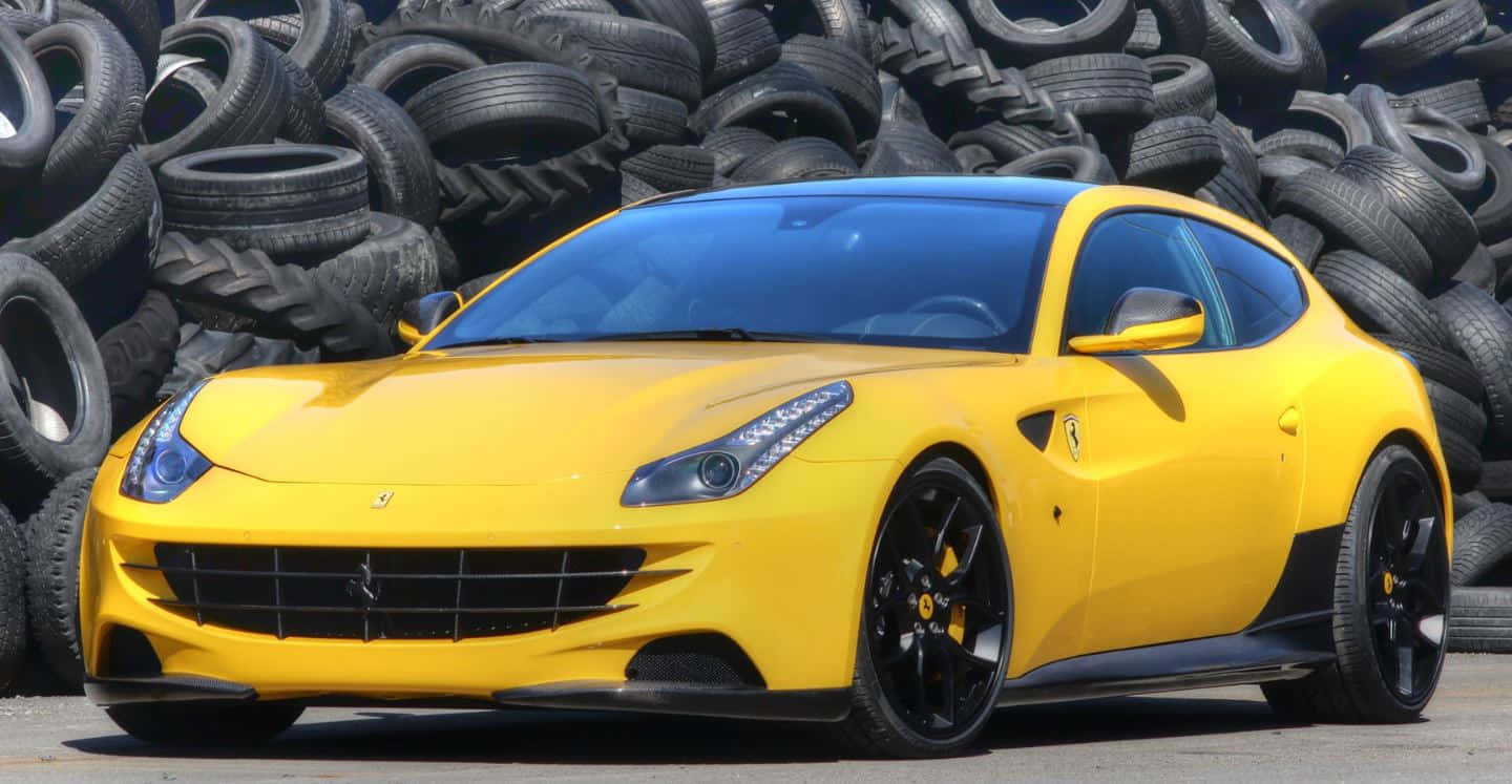 Ferrari Ff 桌布 壁紙 — 一輛充滿活力的黃色法拉利跑車，帶有黑色裝飾，在明亮的日光下大膽地矗立在一堵高聳的輪胎堆疊牆前。