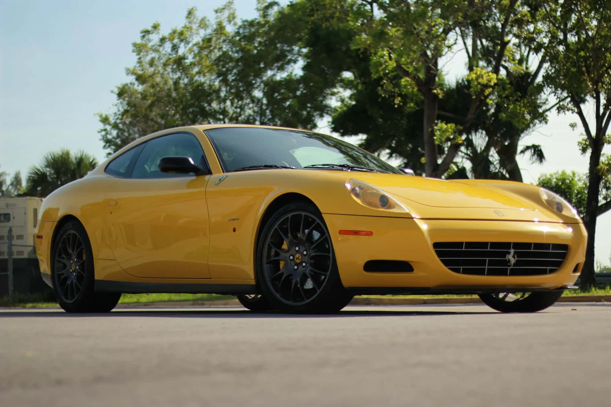 Ferrari 612 Scaglietti 桌布 壁紙 — 一輛充滿活力的黃色法拉利跑車在明亮的陽光下閃閃發光，停在瀝青路上，背景是茂密的綠樹。
