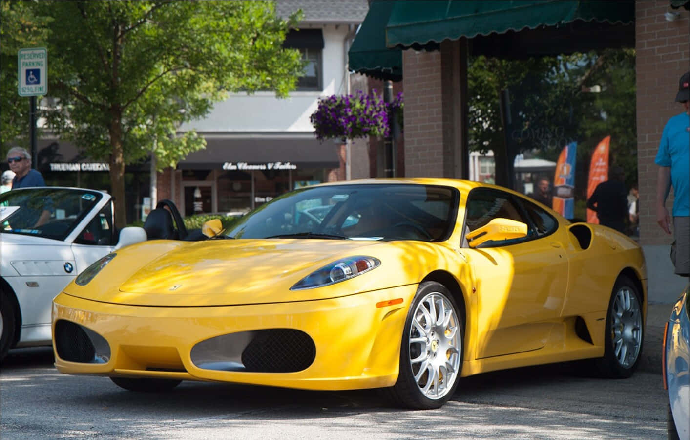 Ferrari F430 桌布 壁紙 — 一輛充滿活力的黃色法拉利跑車在明亮的陽光下閃閃發光，停在綠樹成蔭的城市街道上，背景有行人和商店櫥窗。