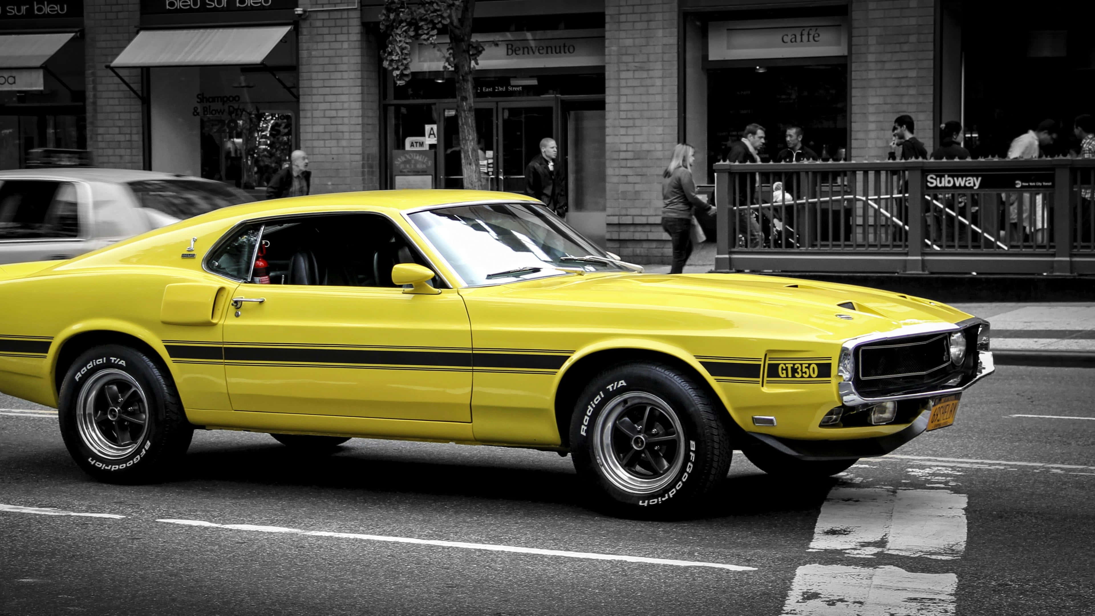 Ford Mustang 桌布 壁紙 — 一輛充滿活力的黃色經典肌肉車在黑白背景的繁華城市街道上行駛。
