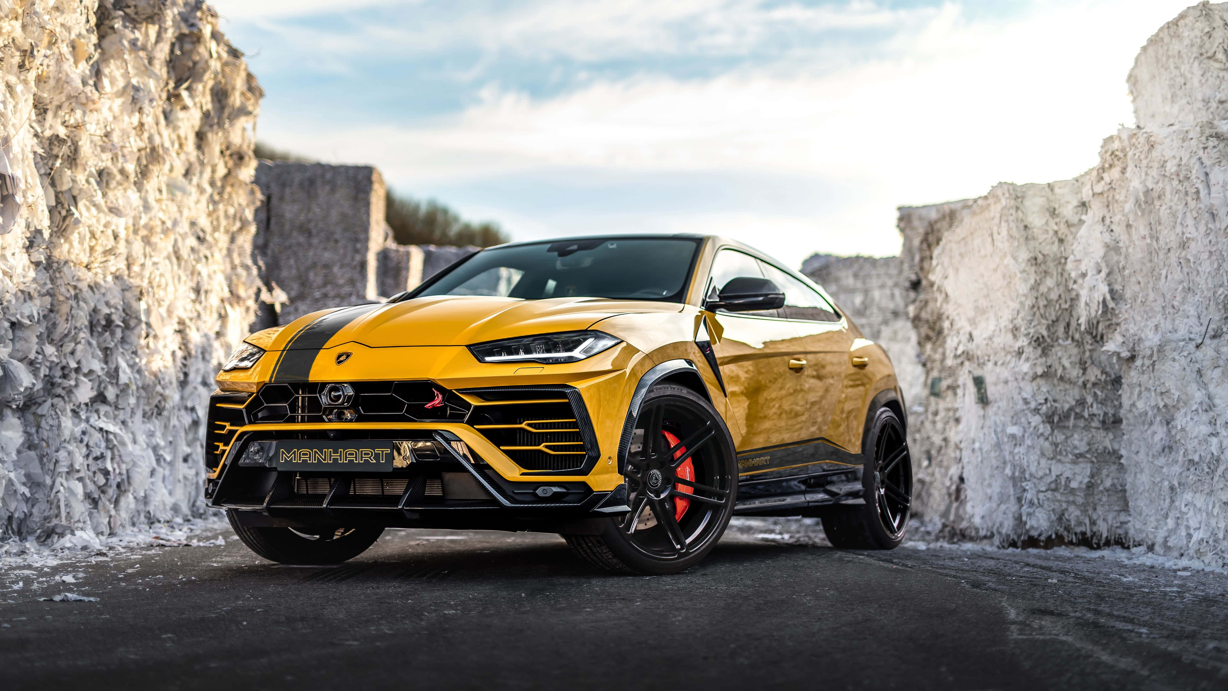 Lamborghini Urus 桌布 壁紙 — 一輛充滿活力的黃色藍寶堅尼 Urus，帶有黑色飾件和紅色制動卡鉗，停泊在聳立的白色石崖之間，天空部分多雲。