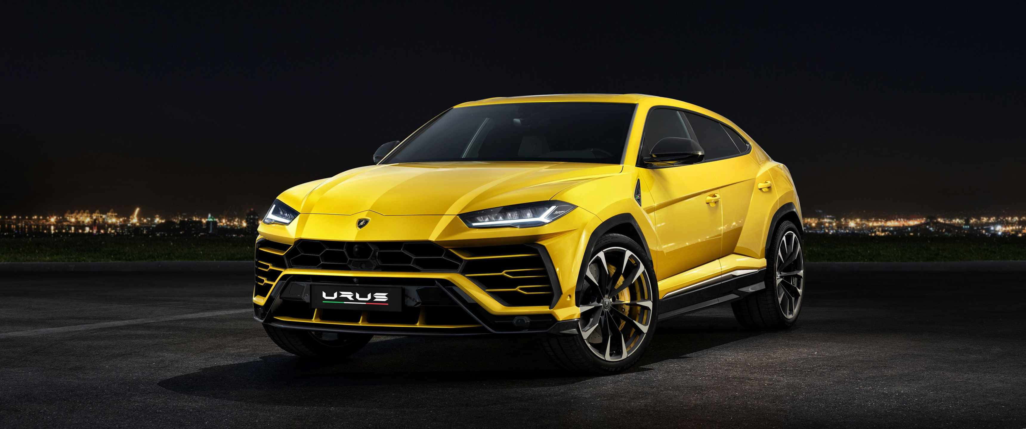 3440x1440p Lamborghini 桌布 壁紙 — 一輛充滿活力的黃色藍寶堅尼 Urus SUV 在夜燈下閃閃發光，遠方城市天際線閃爍。