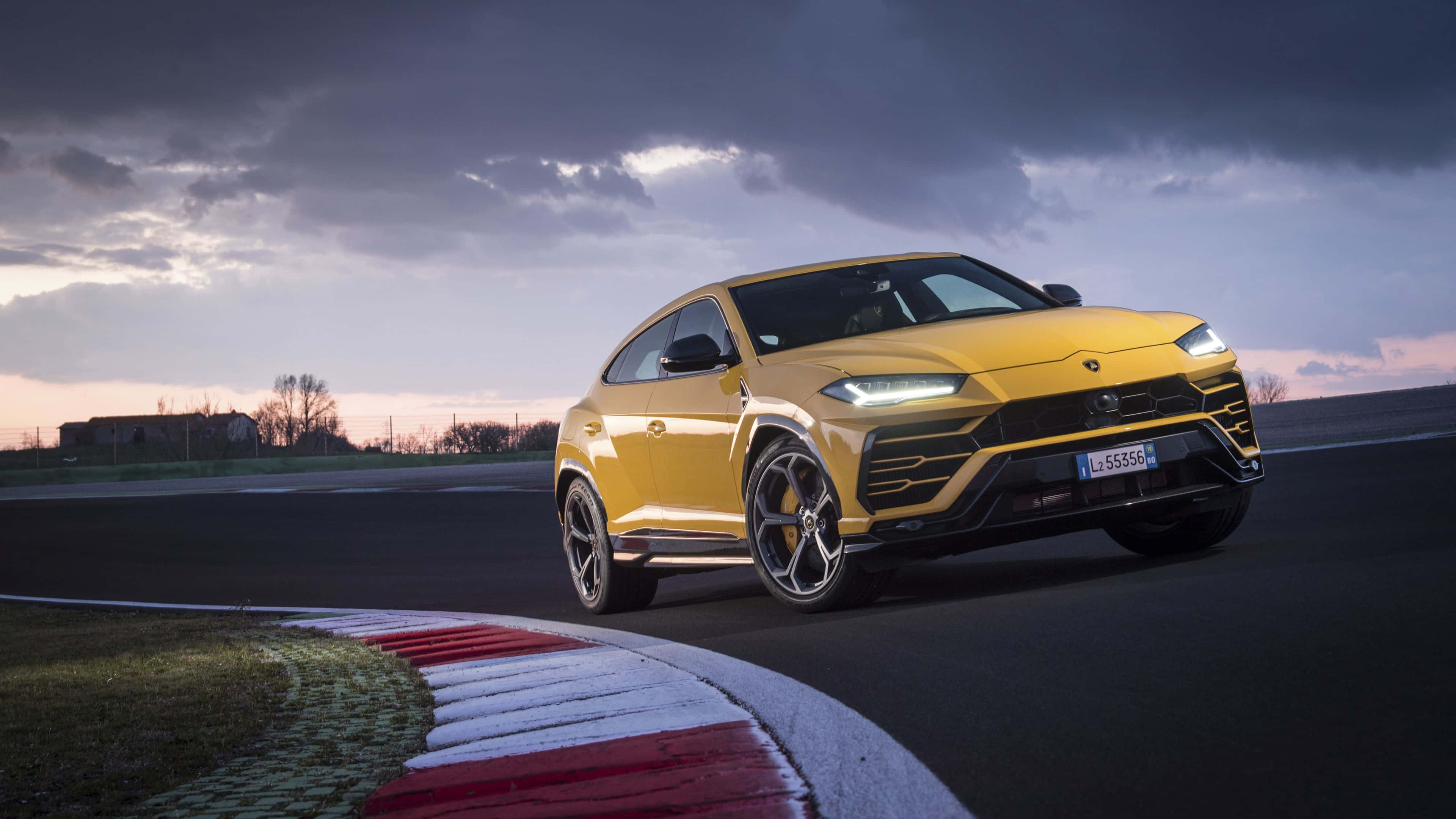 Lamborghini Urus 桌布 壁紙 — 一輛充滿活力的黃色藍寶堅尼 Urus 在戲劇性的暮色天空下於賽道彎角疾馳。
