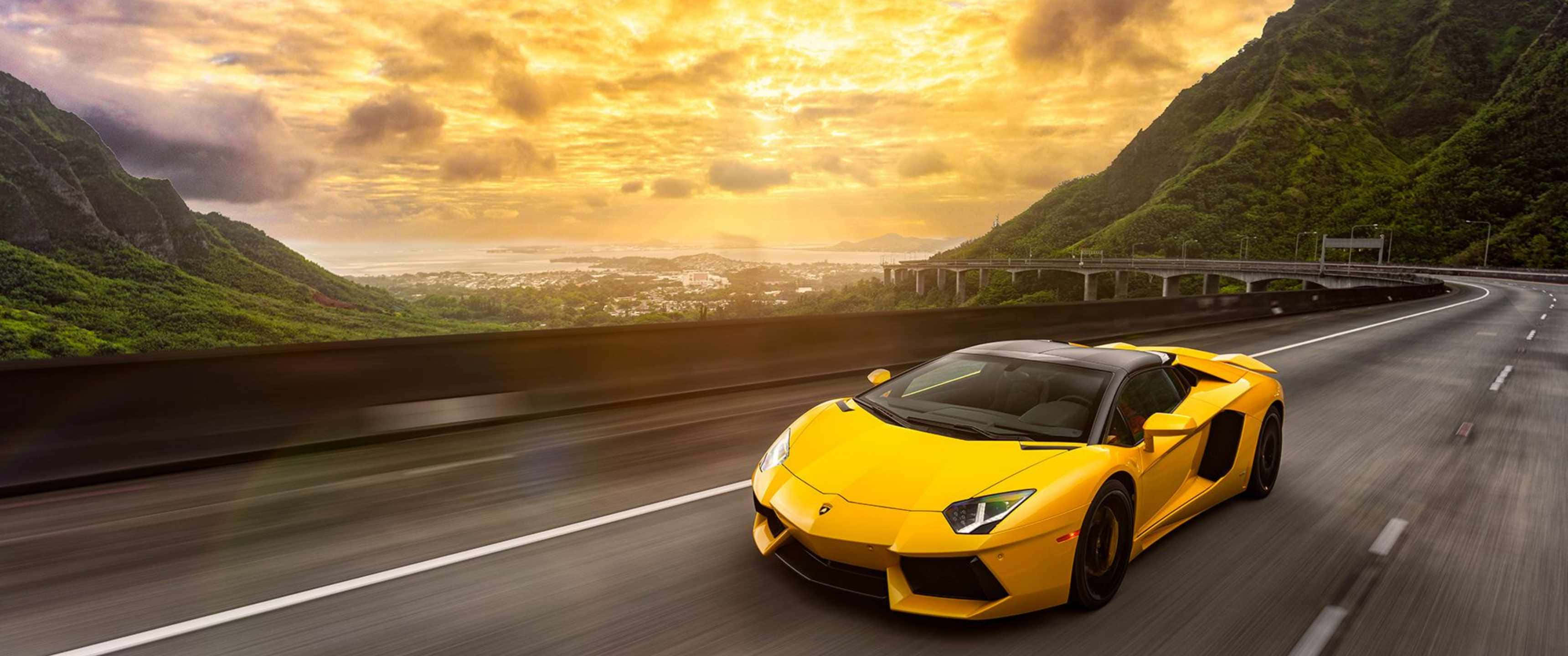 3440x1440p Lamborghini 桌布 壁紙 — 一輛充滿活力的黃色藍寶堅尼在金色日落天空下沿著蜿蜒的山區高速公路飛馳。