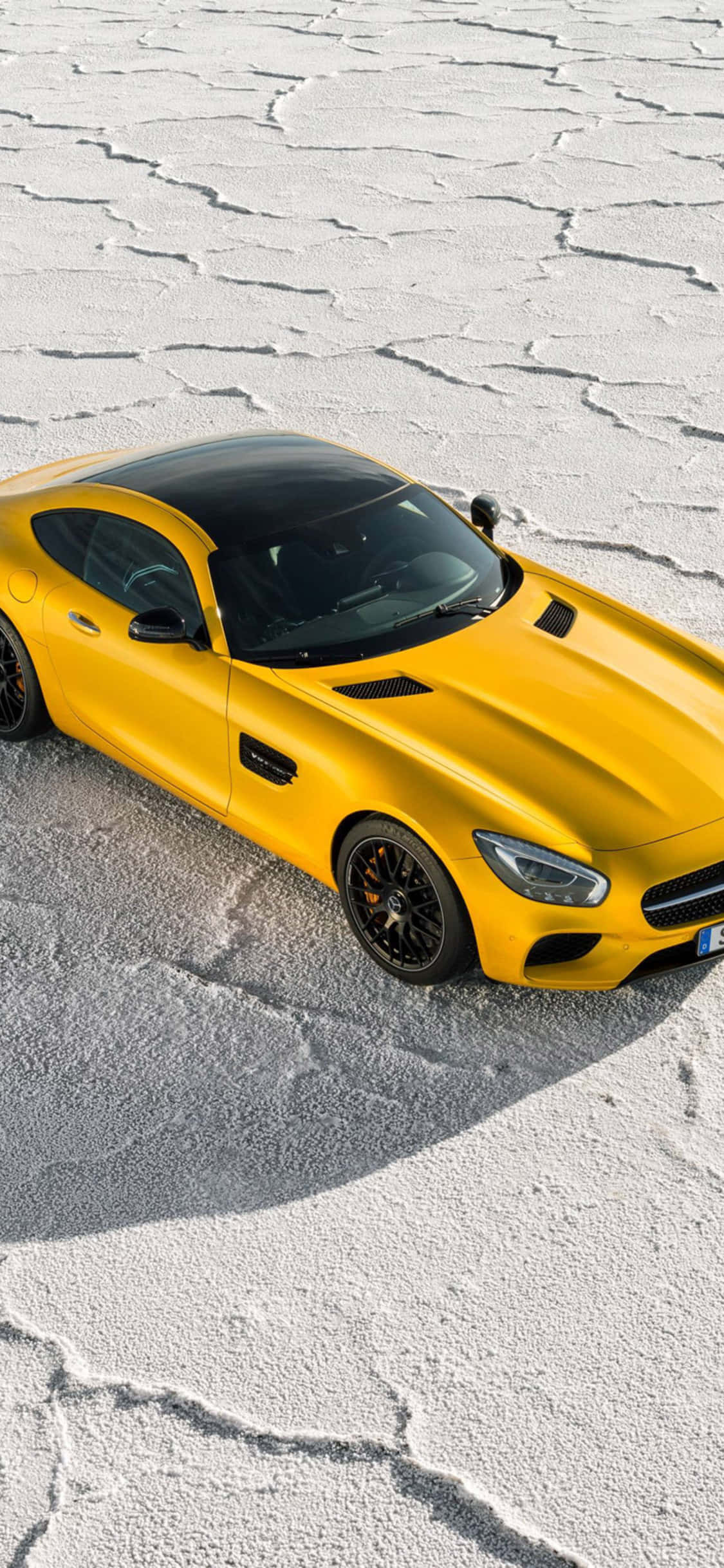 iPhone X Amg Gt-r 桌布 壁紙 — 一輛充滿活力的黃色跑車帶有黑色裝飾，停在龜裂的白色鹽灘上，沐浴在明亮的日光下。
