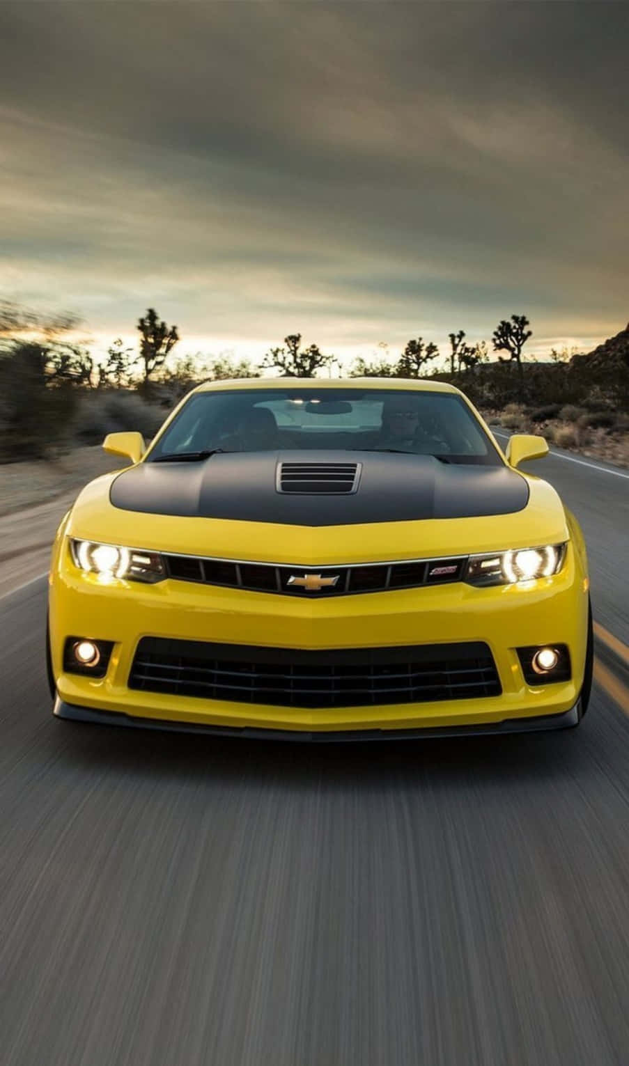 Chevrolet 桌布 壁紙 — 一輛充滿活力的黃色雪佛蘭 Camaro SS 在黃昏時分沿著沙漠公路飛馳，車頭燈閃爍，帶有動態模糊效果。