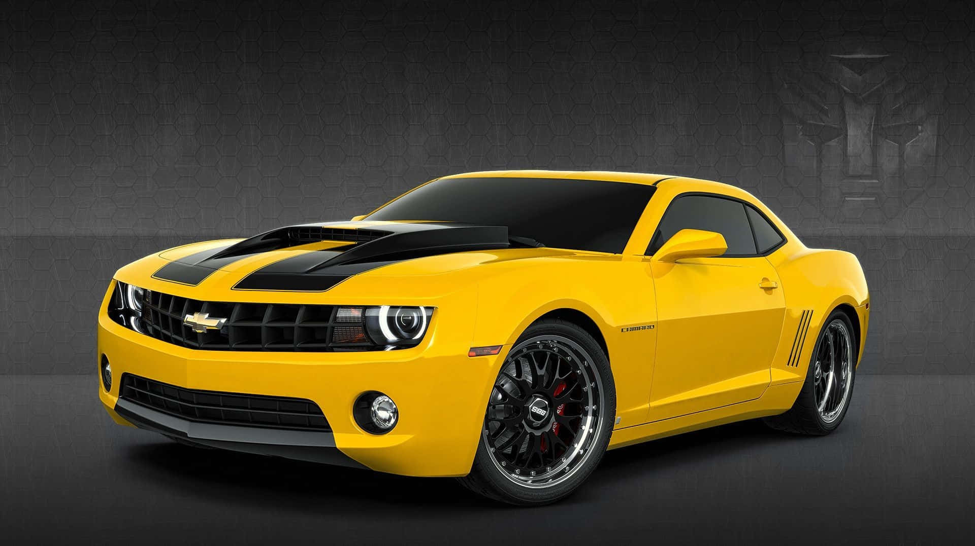 Yellow Car 桌布 壁紙 — 一輛充滿活力的黃色雪佛蘭Camaro，帶有黑色賽車條紋和定制輪轂，在帶有微弱變形金剛標誌的深色紋理牆壁前蓄勢待發。