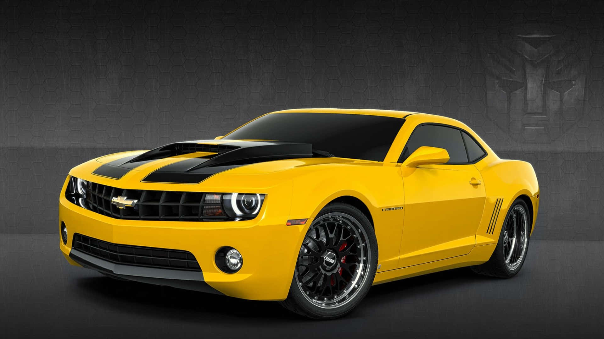 Chevrolet 桌布 壁紙 — 一輛充滿活力的黃色雪佛蘭Camaro，帶有黑色賽車條紋和定制輪圈，在帶有變形金剛標誌的深色紋理牆前擺姿勢。