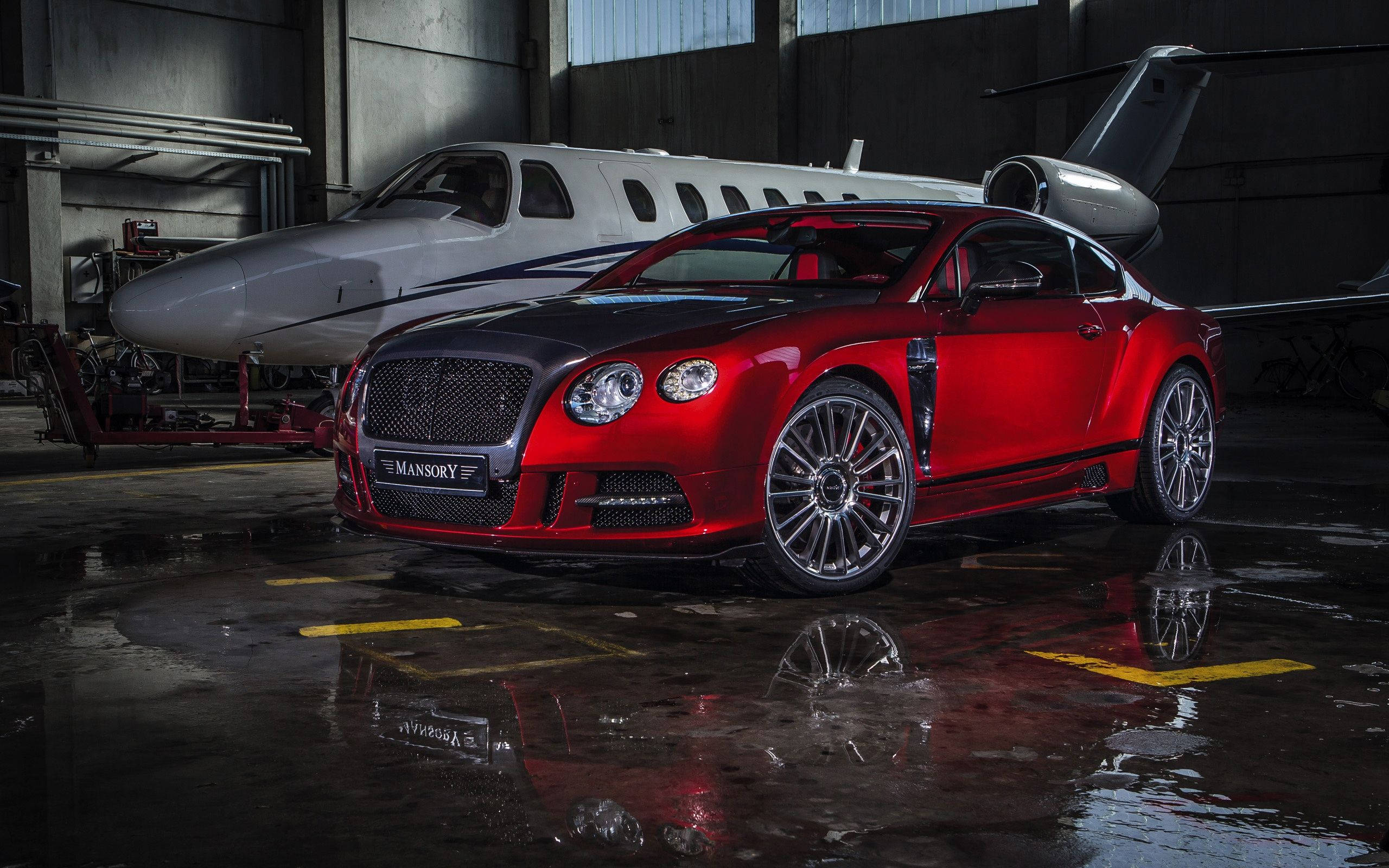 賓利 桌布 壁紙 — 一輛光亮的紅色 Mansory Bentley Continental GT 在濕漉漉的機庫中，於工業燈光下在私人飛機旁閃耀。