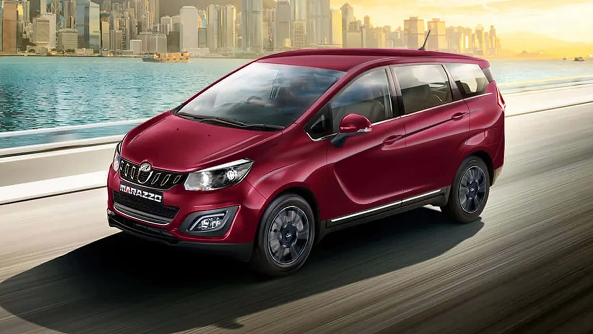 Mahindra Marazzo 桌布 壁紙 — 一輛光亮的紅色 Marazzo 小型貨車沿著海濱高速公路飛馳，現代城市的天際線在金色夕陽中閃耀。