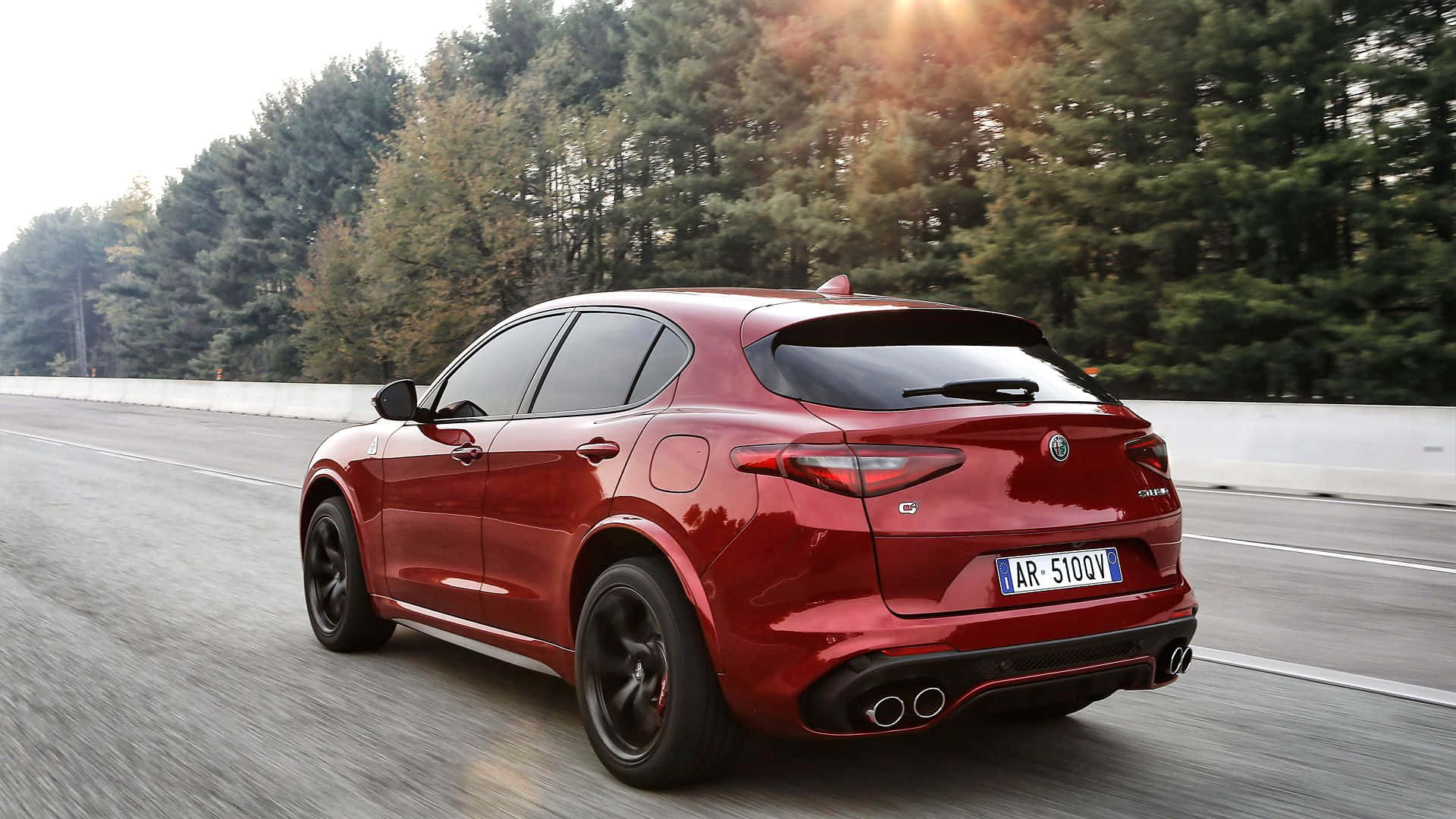 Alfa Romeo Stelvio 桌布 壁紙 — 一輛光亮的紅色Alfa Romeo Stelvio SUV在樹木成行的道路上飛馳，陽光穿過森林樹冠照射下來。