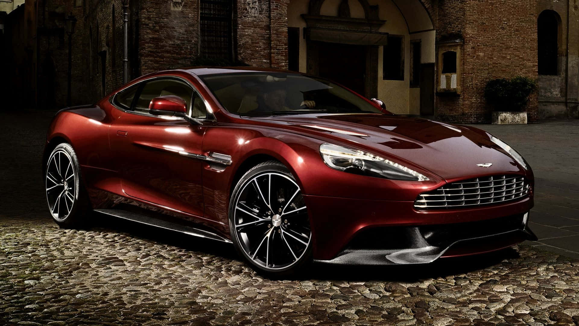 Aston Martin Vanquish 桌布 壁紙 — 一輛光亮的紅色Aston Martin跑車在戲劇性燈光下，在歷史磚砌建築旁的鵝卵石街道上閃閃發光。