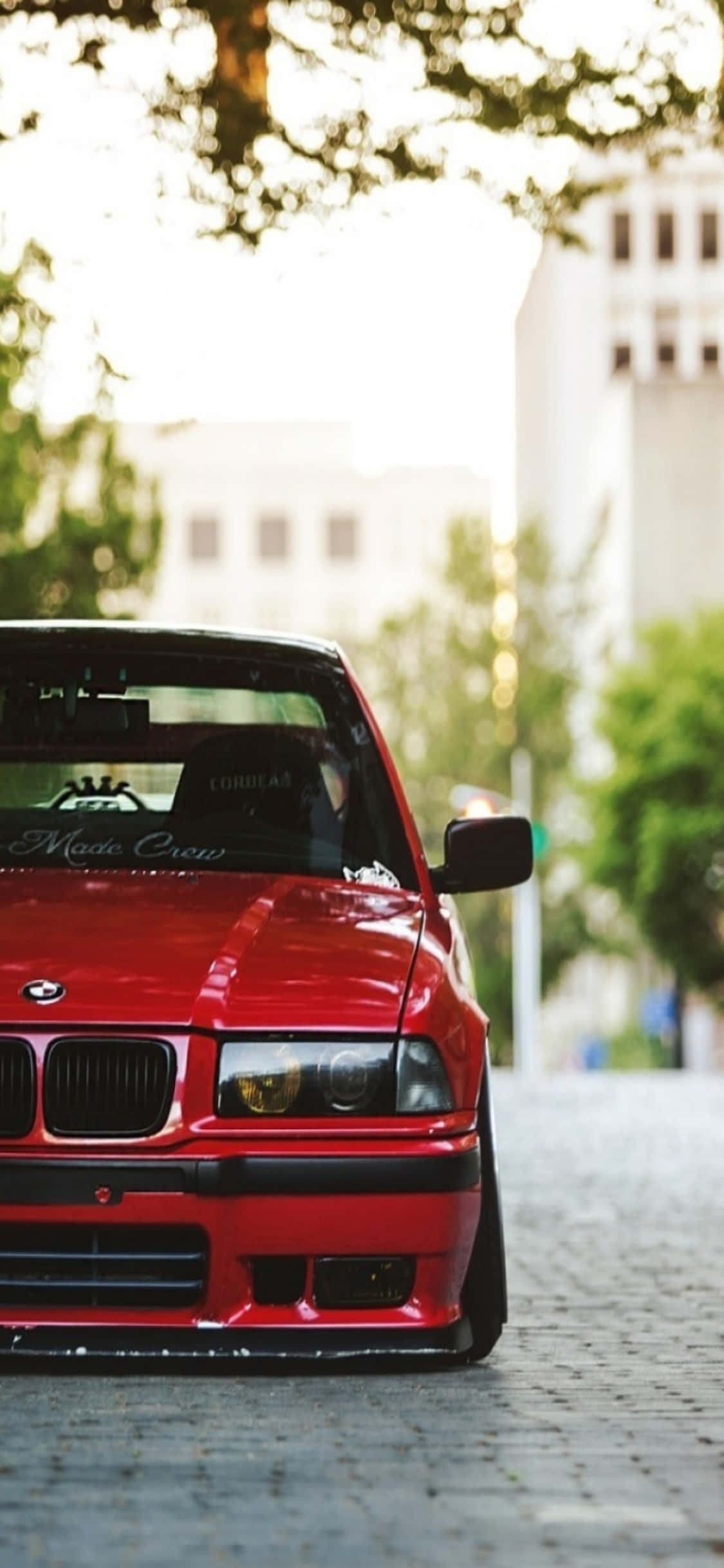 iPhone X Bmw 桌布 壁紙 — 一輛光亮的紅色BMW E30跑車停在鵝卵石街道上，斑駁的陽光下，背景有模糊的綠色樹木和建築物。