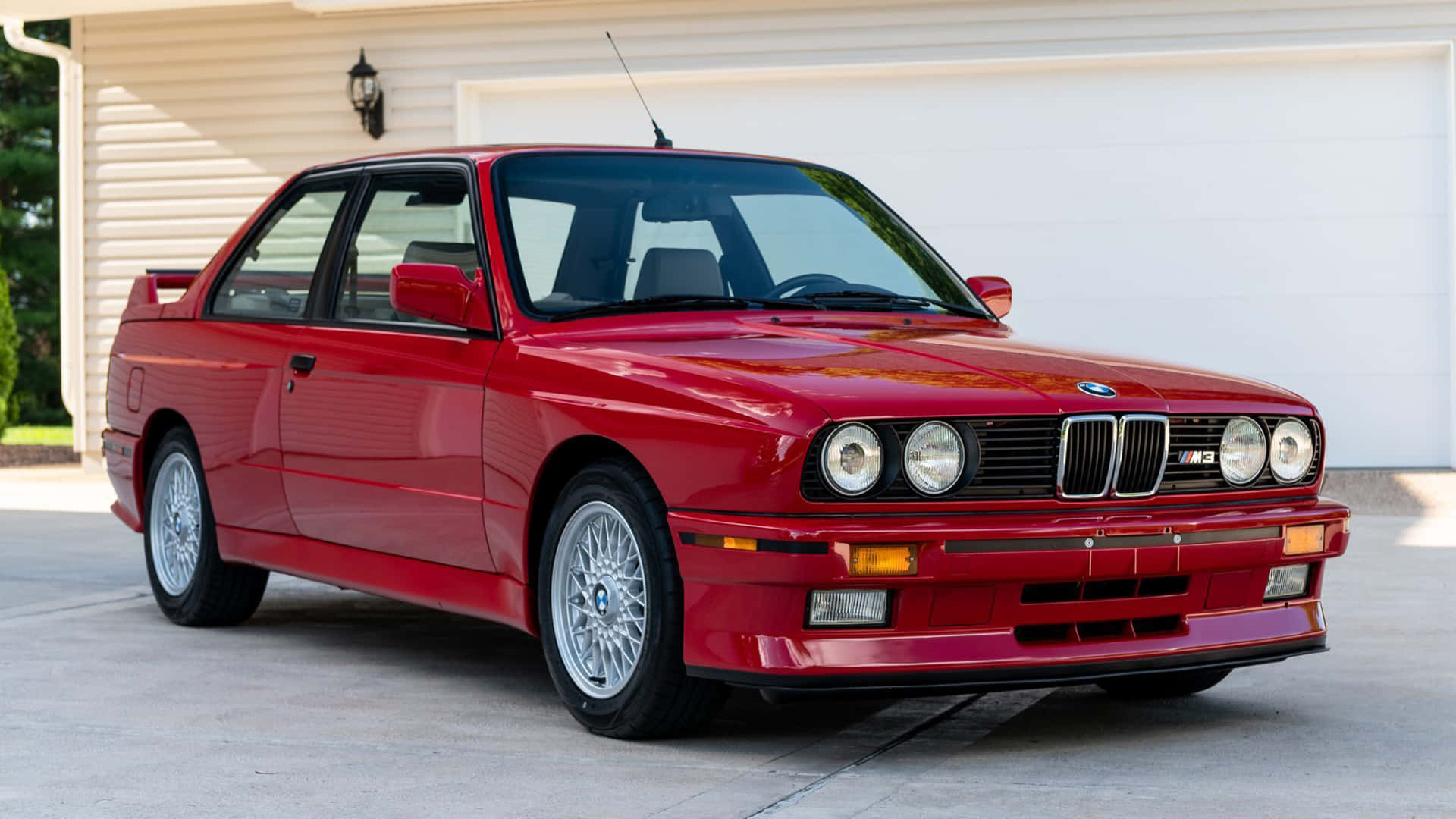 BMW 動圖 桌布 壁紙 — 一輛光亮的紅色BMW M3跑車停放在白色車庫前的混凝土車道上。