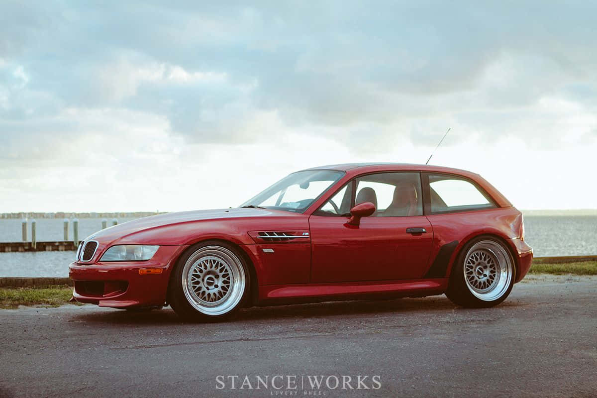 BMW 動圖 桌布 壁紙 — 一輛光亮的紅色BMW Z3 M Coupe配備客製輪圈，停泊在多雲天空下的水邊柏油路上。