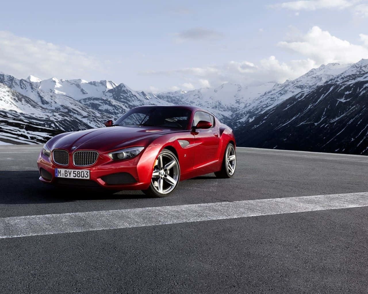 Bmw Z4 桌布 壁紙 — 一輛光亮的紅色BMW Z4跑車停在山路上，背景是部分多雲的天空下的積雪山峰。