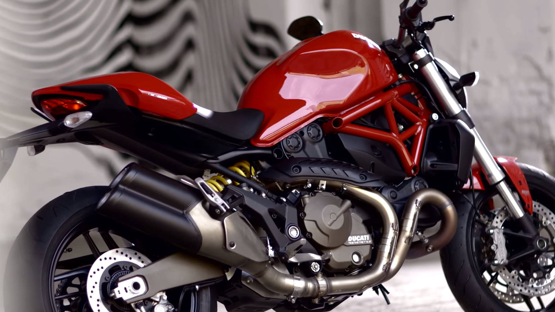 杜卡迪 桌布 壁紙 — 一輛光亮的紅色Ducati機車，露出引擎和鍍鉻排氣管，在波浪形灰色背景上閃閃發光。