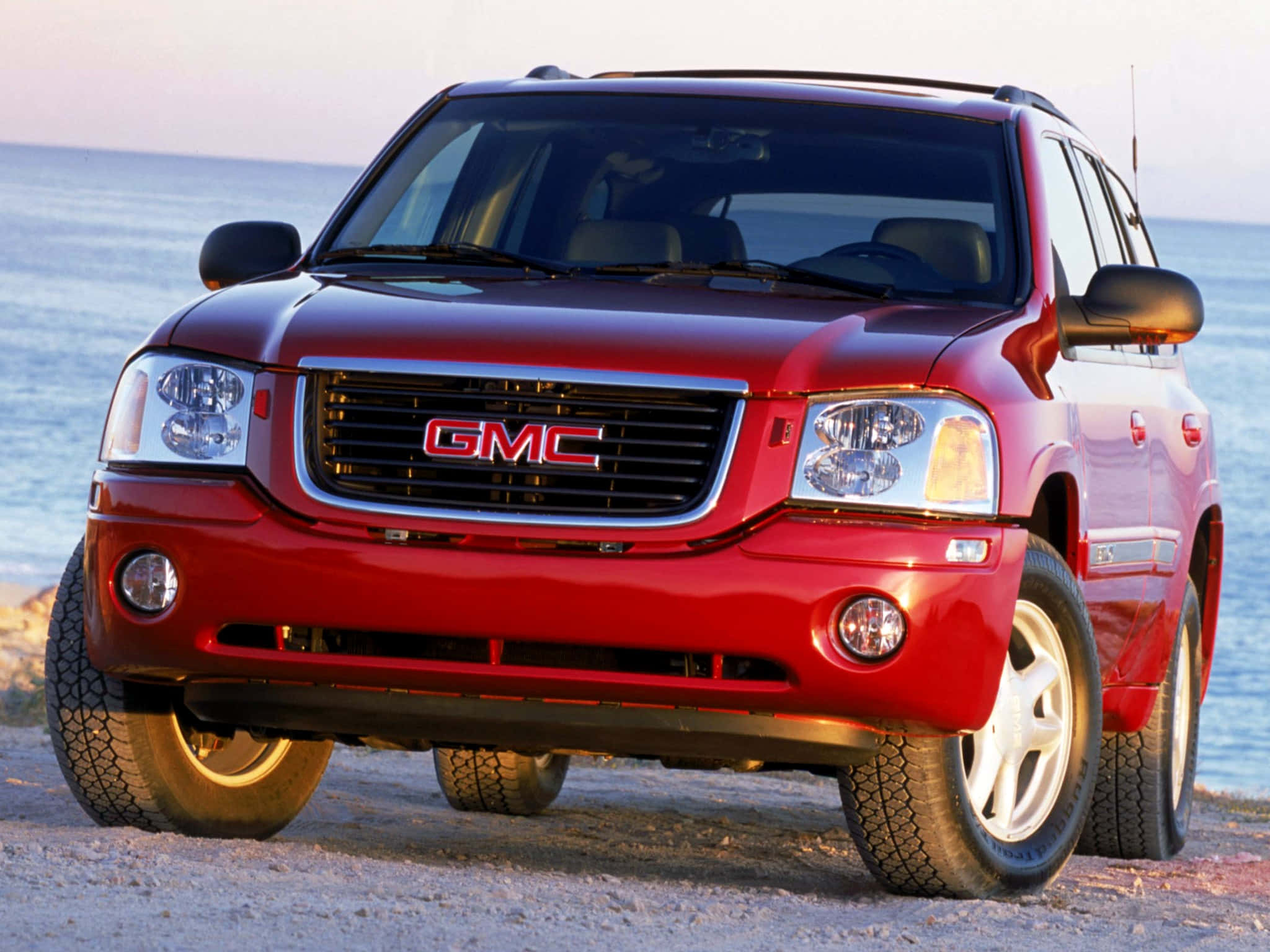 Gmc Envoy 桌布 壁紙 — 一輛光亮的紅色GMC SUV停泊在沙灘上，背景是日落時分的海浪。