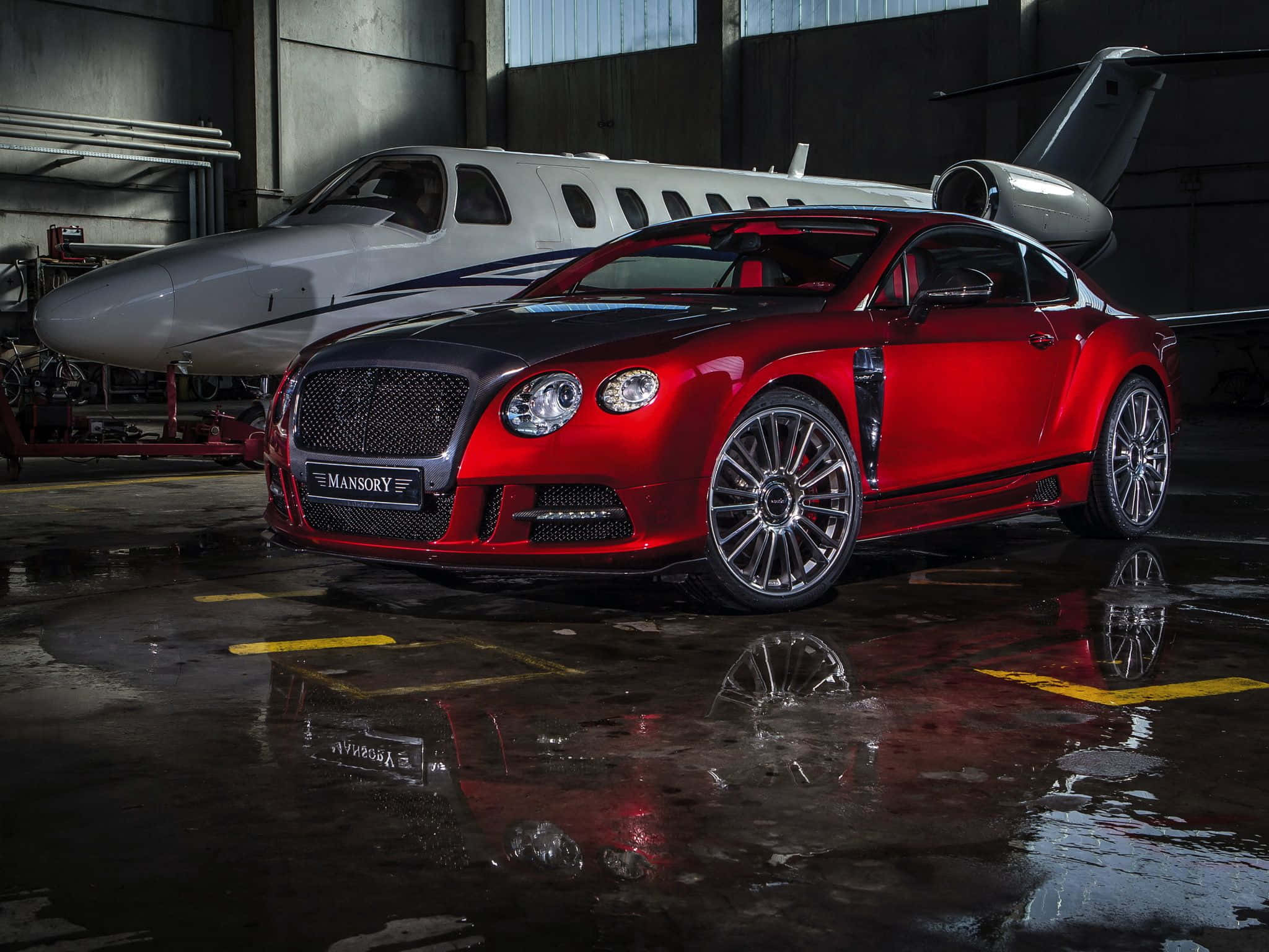 賓利 桌布 壁紙 — 一輛光亮的紅色Mansory Bentley Continental GT轎跑車在潮濕的機庫中，在工業燈光下閃耀，旁邊是一架私人飛機。