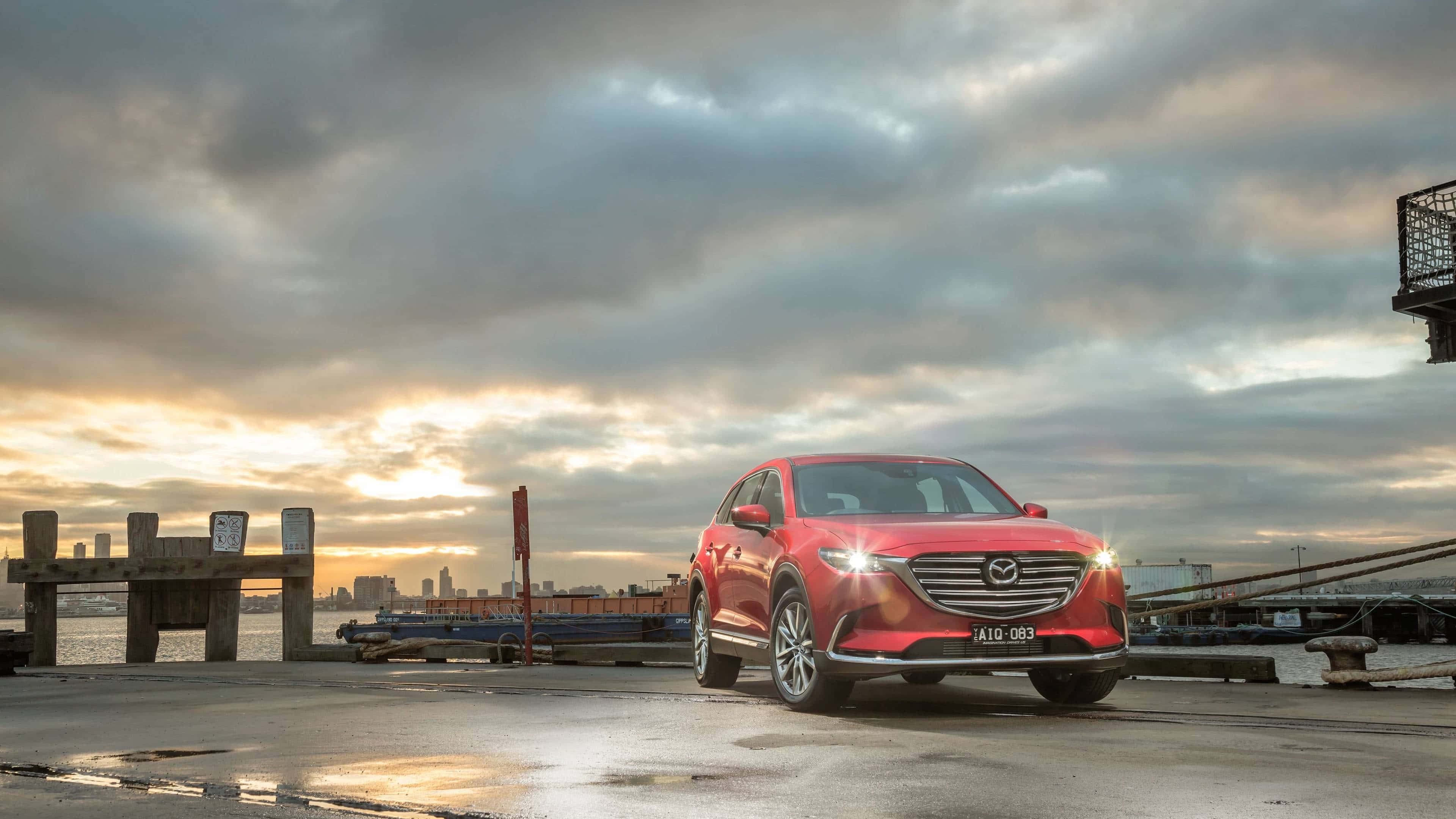 Mazda Cx-9 桌布 壁紙 — 一輛光亮的紅色Mazda CX-9 SUV在戲劇性的日落天空下，在水邊碼頭旁閃閃發光，遠處是城市天際線。