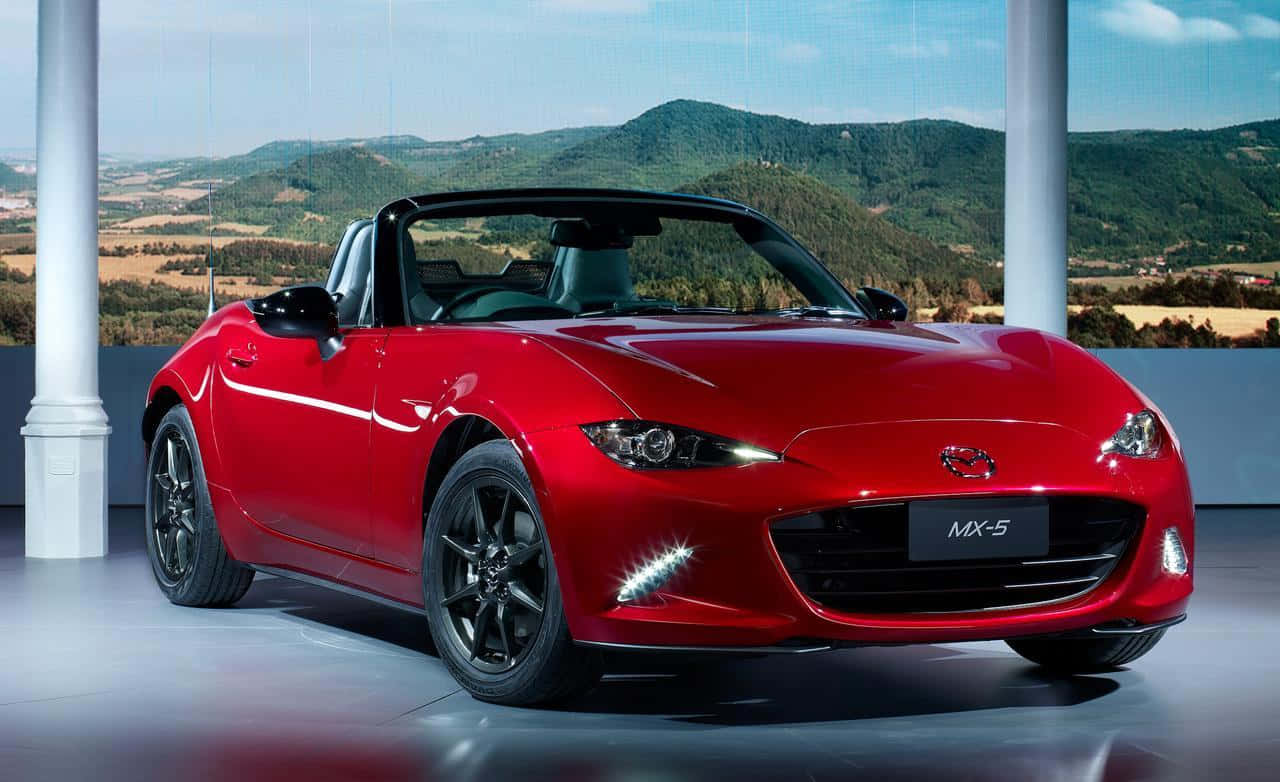 Miata 桌布 壁紙 — 一輛光亮的紅色Mazda MX-5 Miata敞篷車在攝影棚燈光下閃閃發光，背後巨大的柱子之間可以看到風景如畫的山區景色。