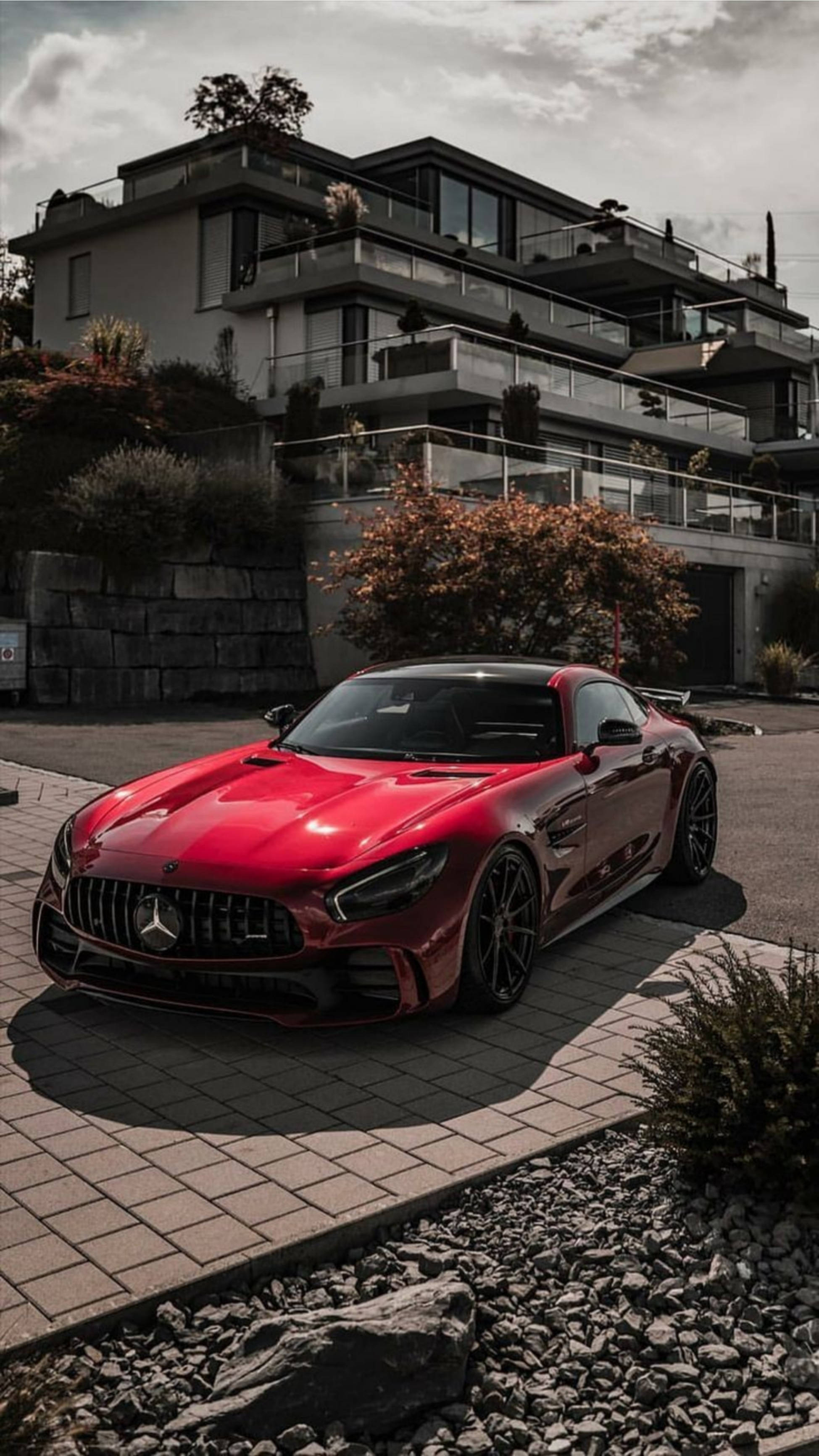 Mercedes 桌布 壁紙 — 一輛光亮的紅色Mercedes-AMG GT停泊在石頭車道上，前方是帶有陽台和秋葉的現代豪華住宅。