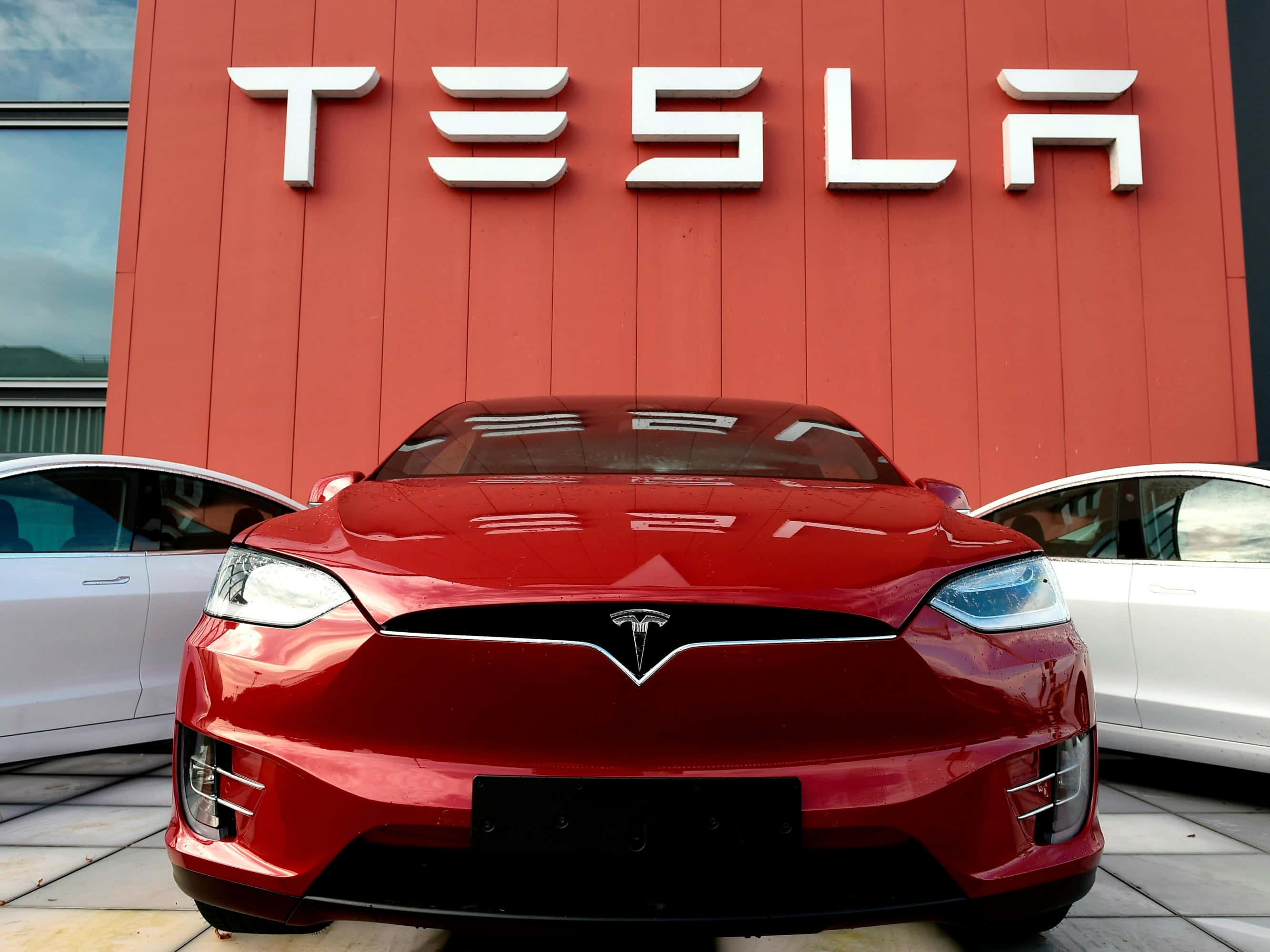 特斯拉 桌布 壁紙 — 一輛光亮的紅色Tesla Model X面向前方，前面是一座大膽的紅色建築，上面有大型白色Tesla標誌。