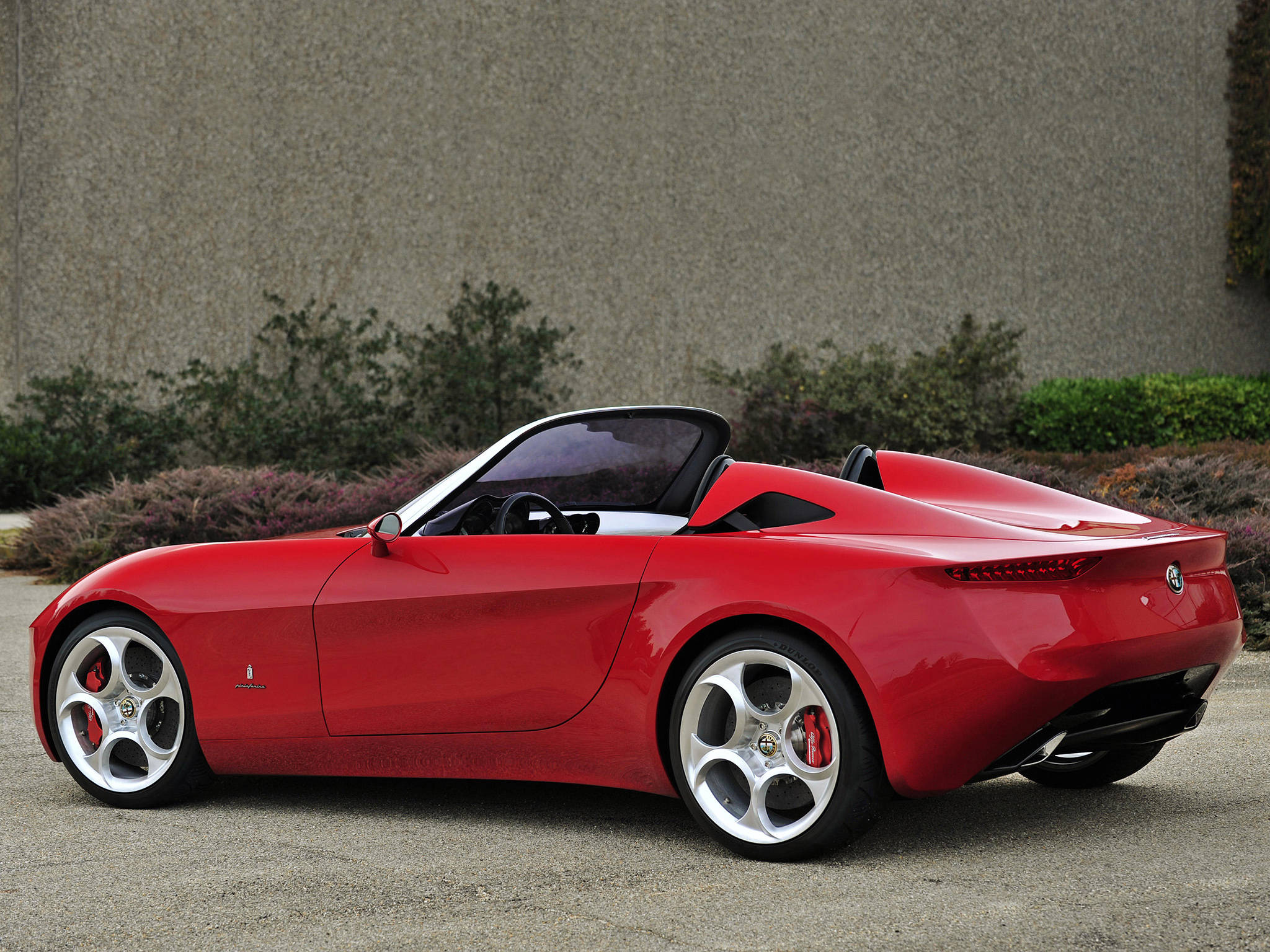 alfa romeo iPhone 桌布 壁紙 — 一輛光亮的紅色愛快羅密歐Spider敞篷車停在碎石上，後面有紋理牆壁和灌木。