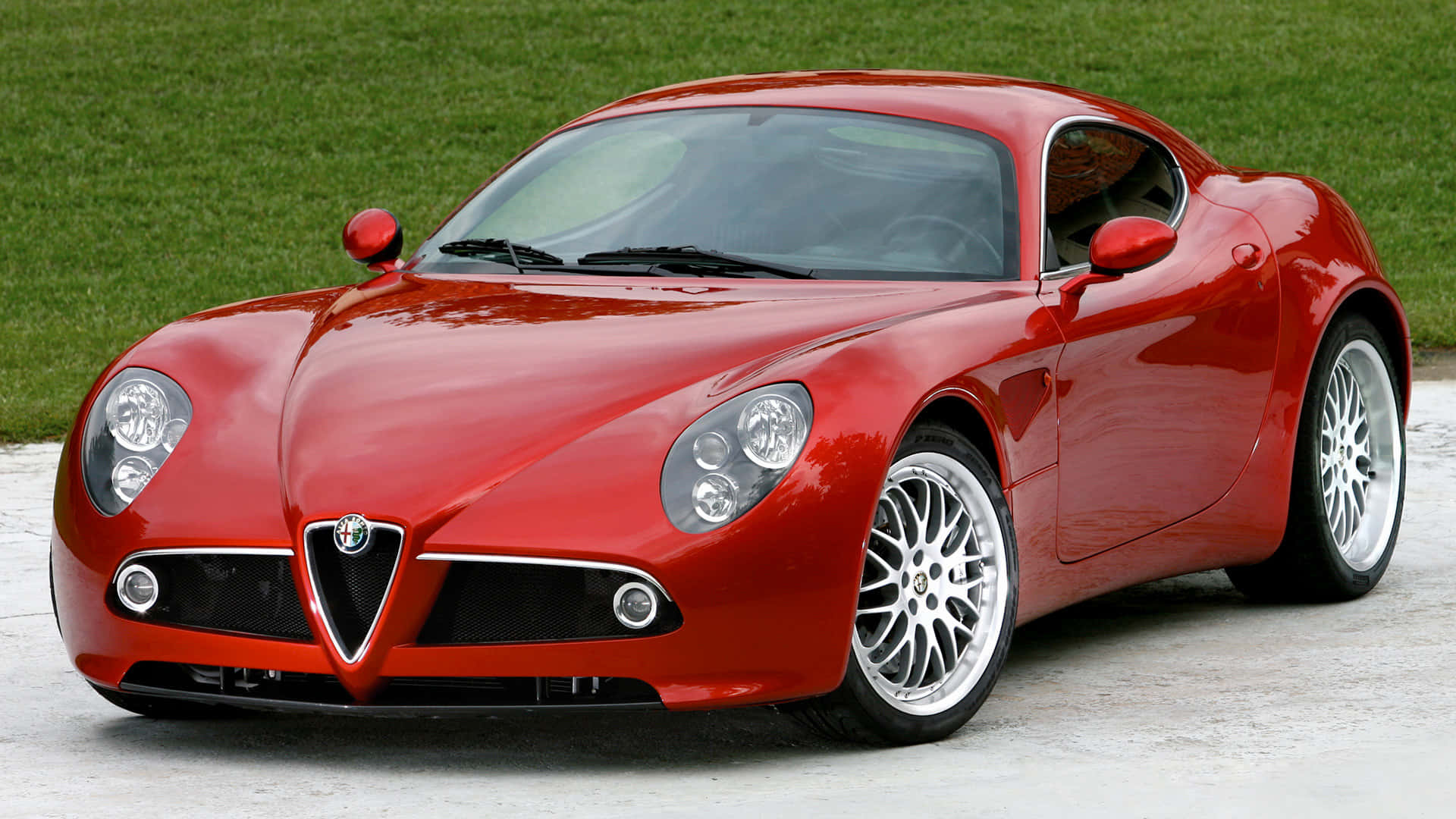 Alfa Romeo 8c Competizione 桌布 壁紙 — 一輛光亮的紅色愛快羅密歐跑車在日光下閃耀，停在混凝土表面上，後面有綠色草地。