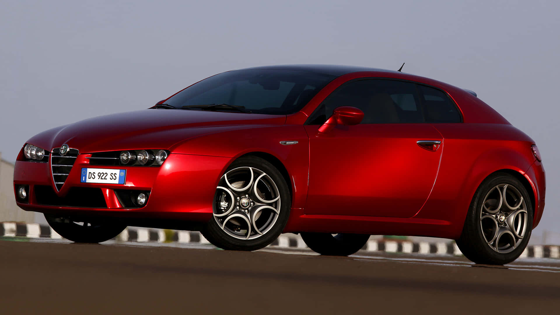 Alfa Romeo Brera 桌布 壁紙 — 一輛光亮的紅色愛快羅密歐跑車在晴朗的天空下精準而有力地沿著賽道滑行。