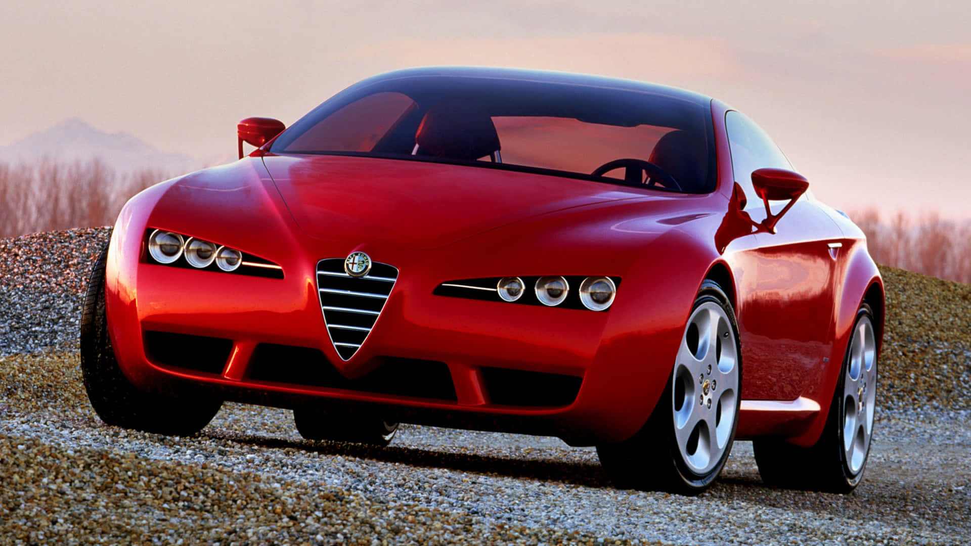 Alfa Romeo Brera 桌布 壁紙 — 一輛光亮的紅色愛快羅密歐跑車在碎石路上閃耀，背景是柔和的日落天空和遠山。