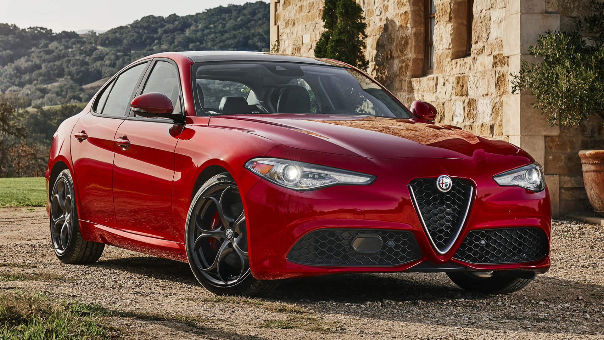 Alfa Romeo Giulia 桌布 壁紙 — 一輛光亮的紅色愛快羅密歐轎車停在碎石路上，旁邊是石頭建築，背景有山丘。