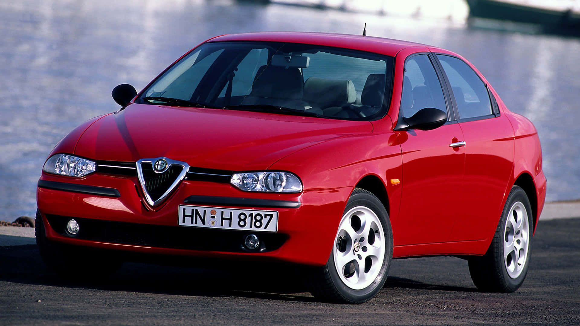 Alfa Romeo 156 桌布 壁紙 — 一輛光亮的紅色愛快羅密歐轎車停泊在河道旁，在明亮的日光下，拋光表面閃爍著倒影。