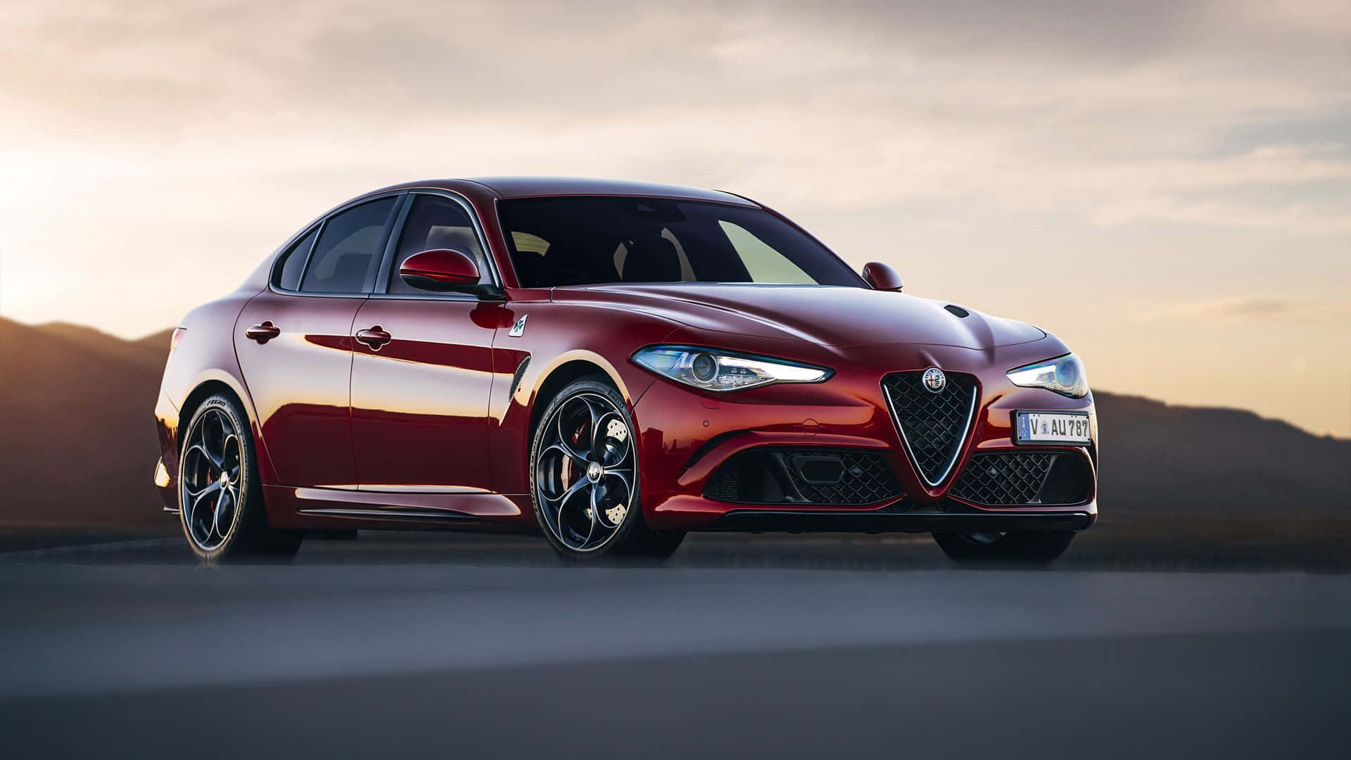 Alfa Romeo Stelvio 桌布 壁紙 — 一輛光亮的紅色愛快羅密歐轎車在金色日落光線下閃耀，行駛在沙漠公路上，遠方有山丘。