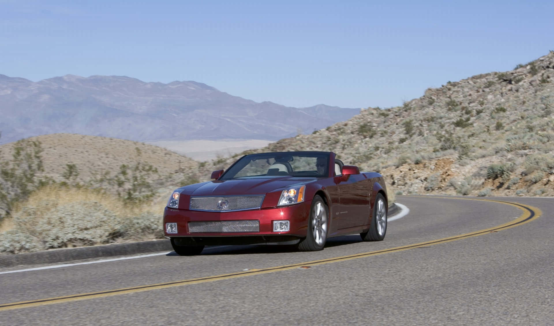 Cadillac Xlr 桌布 壁紙 — 一輛光亮的紅色敞篷跑車在蜿蜒的沙漠公路上行駛，遠山在清澈的藍天下。